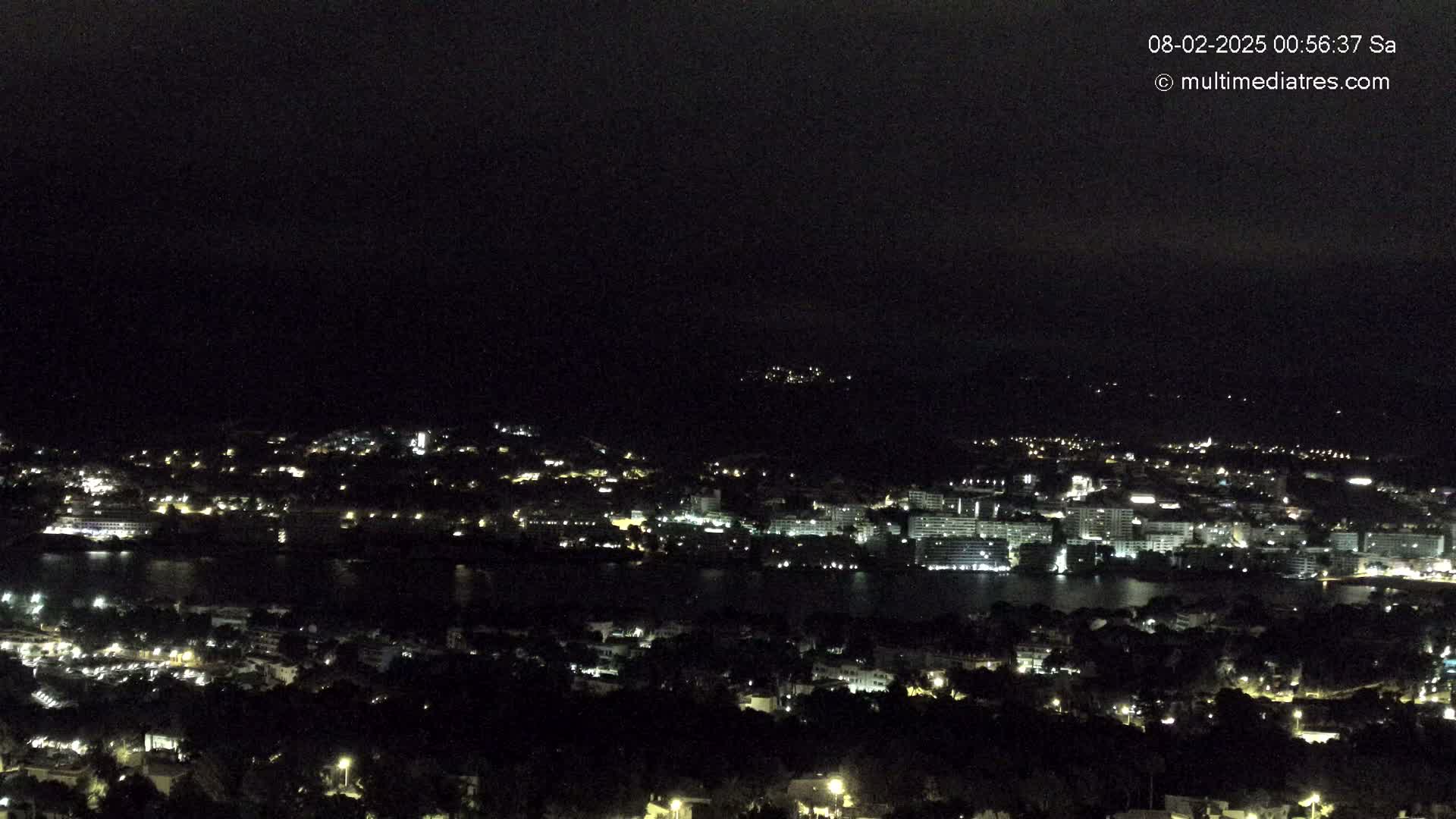 A nighttime view of a coastal town illuminated with scattered lights, under a dark, cloudy sky.
