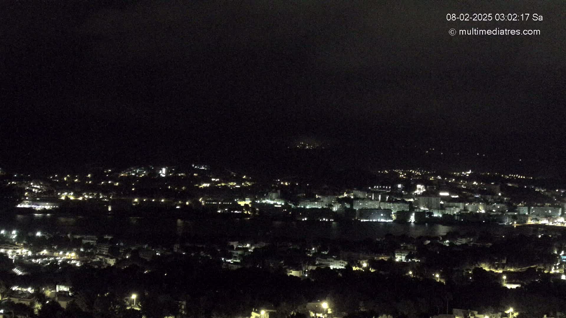 A nighttime view of a city illuminated by scattered lights, situated beside a dark body of water under a mostly cloudy sky.