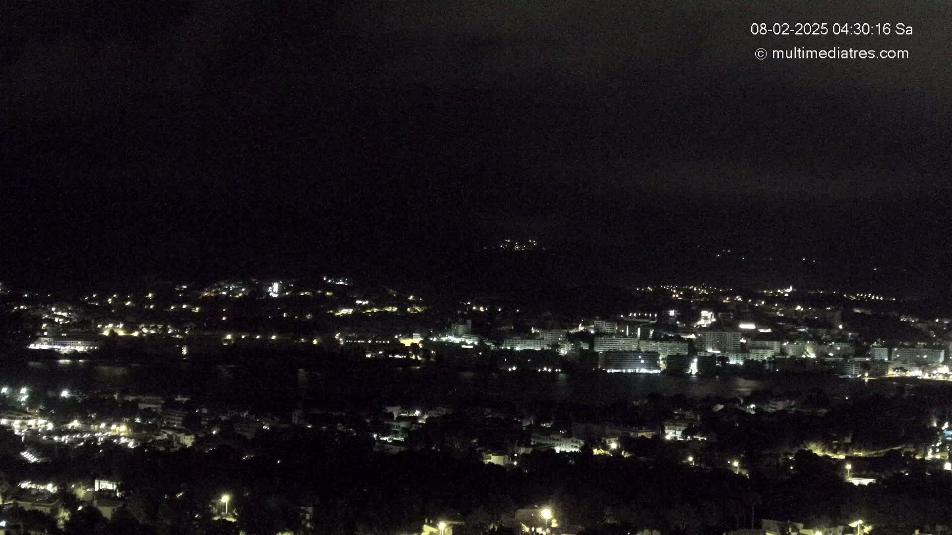 A nighttime, long-range view of a coastal town with scattered lights, situated near a body of water under a dark, cloudy sky.