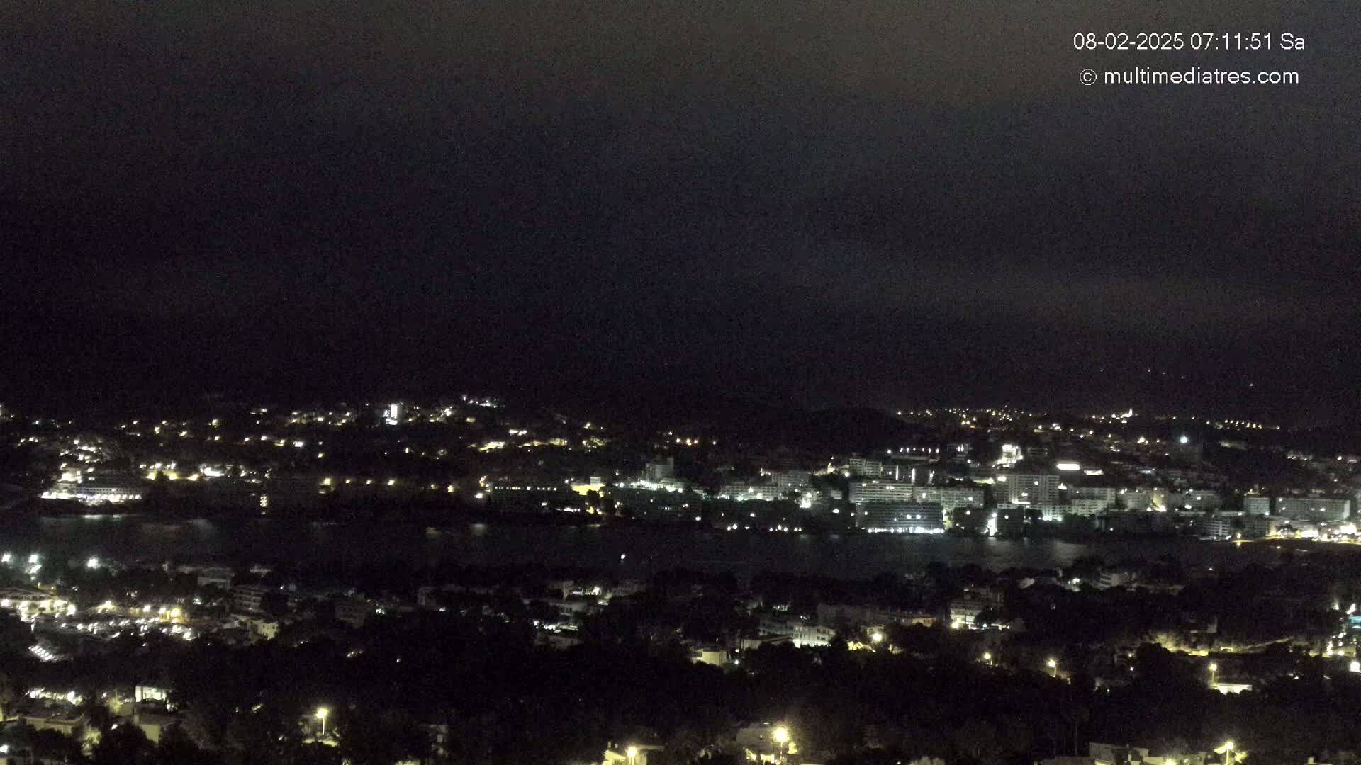 A nighttime, high-angle view of a coastal town with scattered lights, a dark body of water, and a mostly cloudy sky.