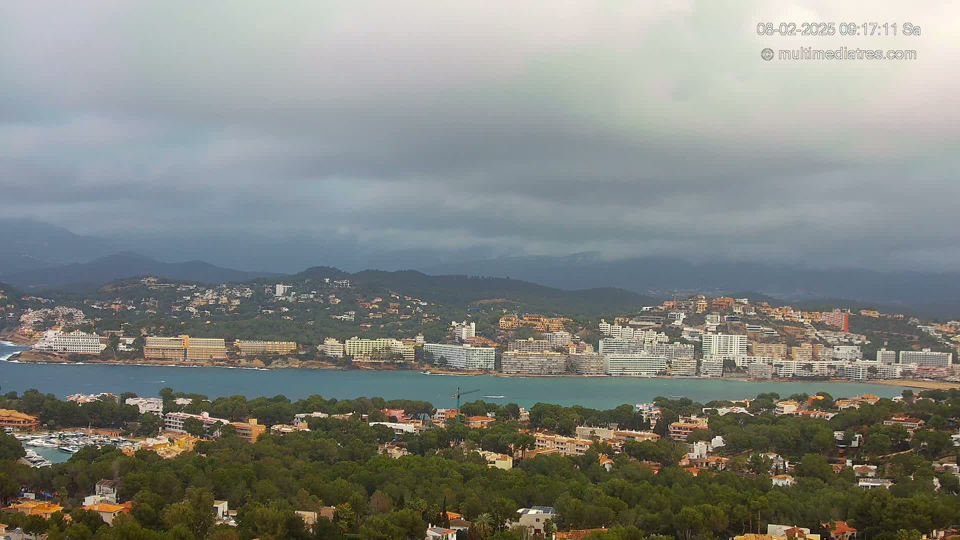 A coastal town nestled among green hills and a calm bay under a cloudy sky.