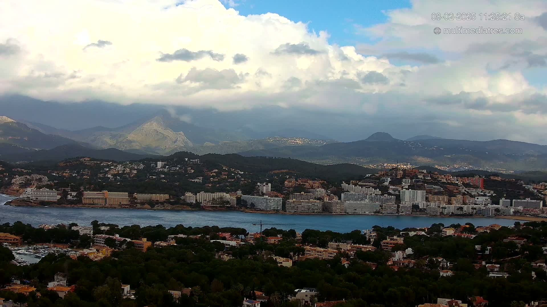 A coastal town nestled between a calm bay and a mountain range under a partly cloudy sky.