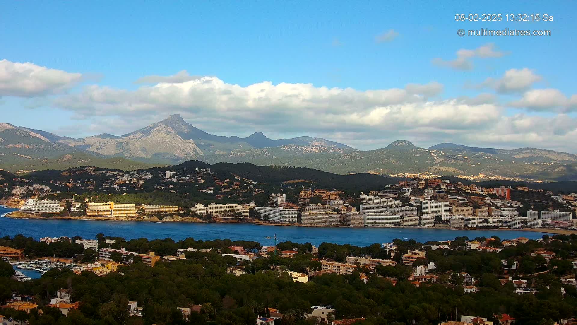 A coastal town with hotels and buildings nestled between a body of water and a mountain range under a partly cloudy blue sky.