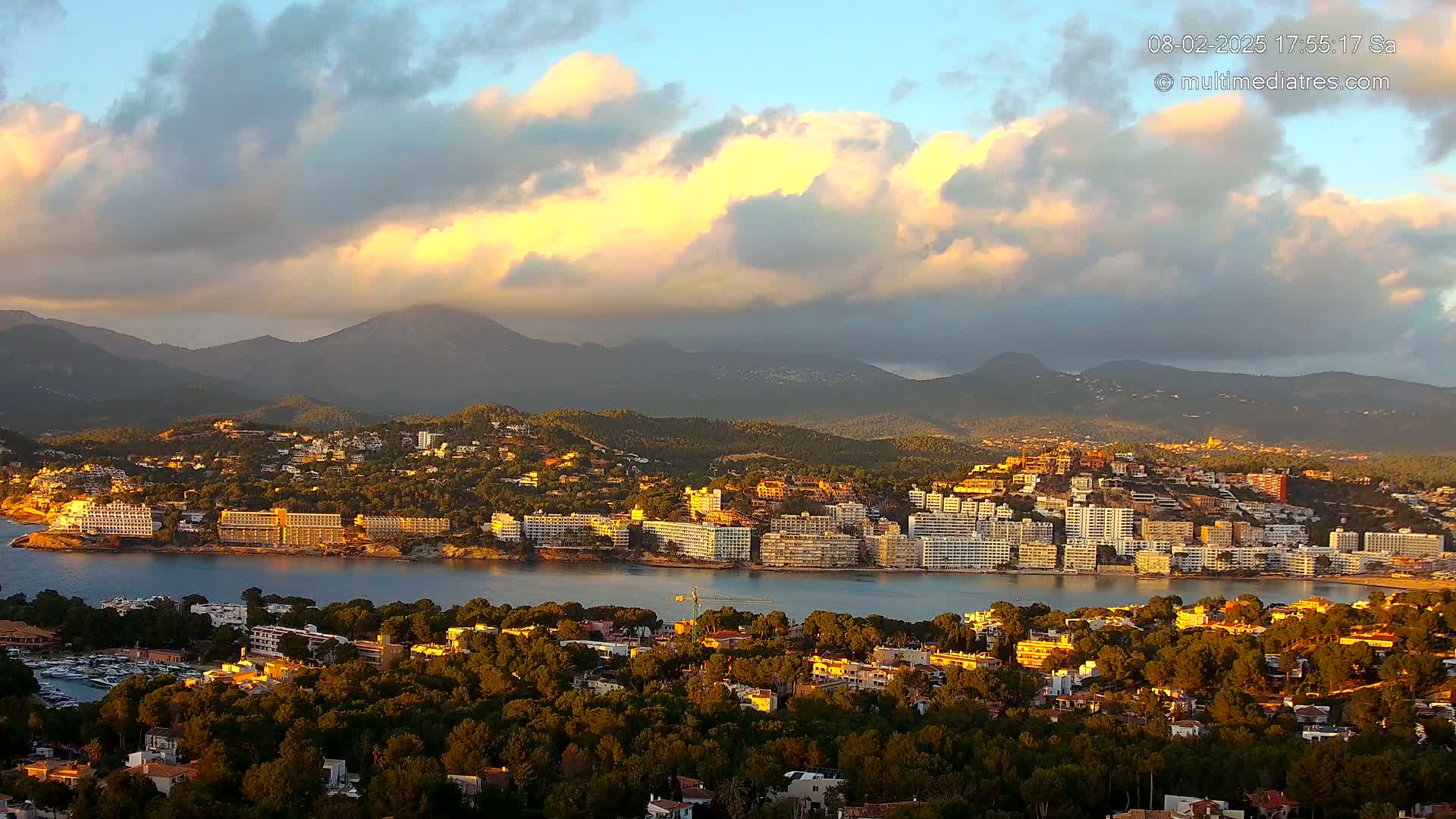 A coastal town nestled among green hills and mountains, with a calm bay and a partly cloudy sunset sky.