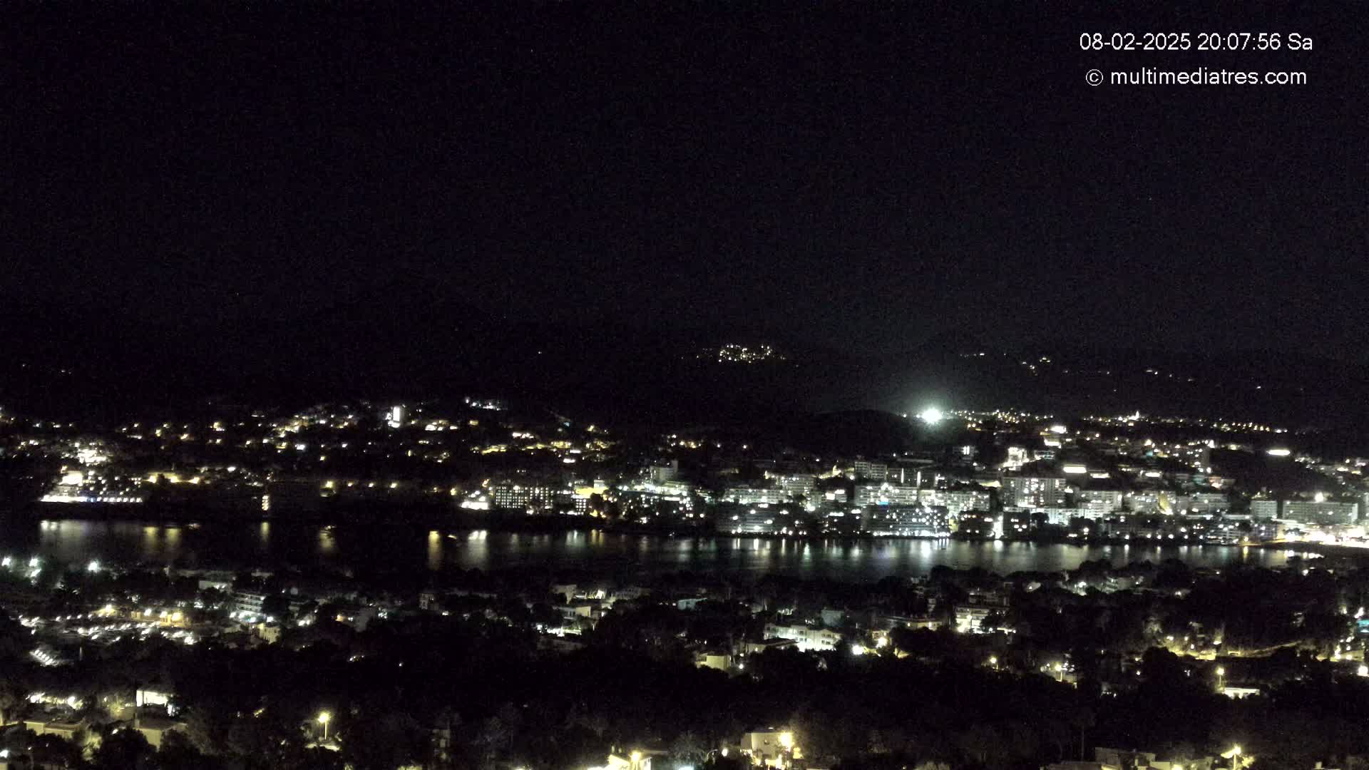 A nighttime, high-angle view shows a city illuminated by lights surrounding a body of dark water, with hills visible in the background under a clear, dark sky.