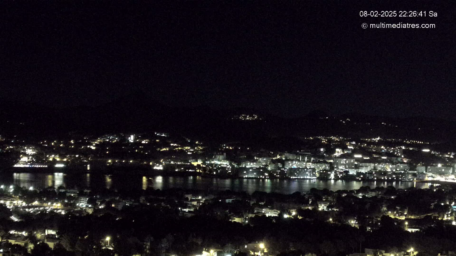 A nighttime view shows a city by a body of water, illuminated by scattered lights, under a clear, dark sky.