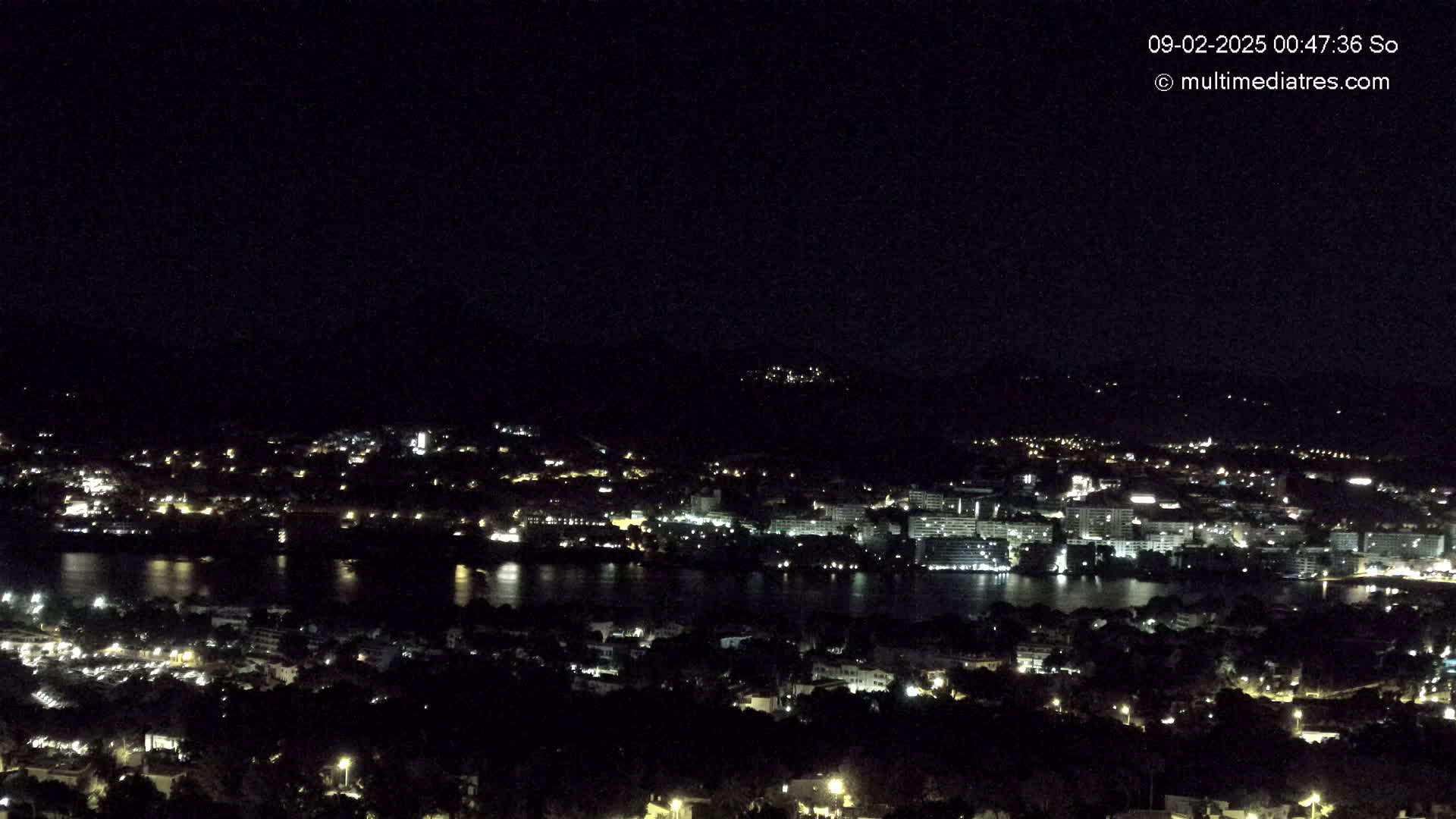 A nighttime view of a coastal city illuminated with many lights, spread across hills and along a body of water under a clear, dark sky.