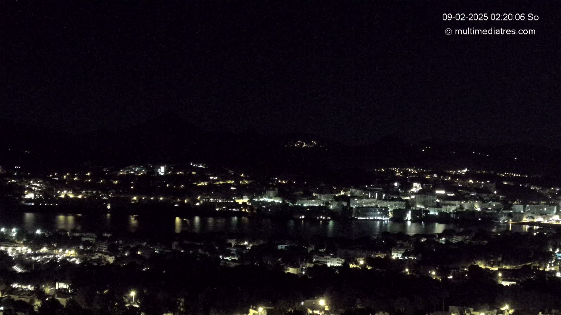 A nighttime, high-angle view of a coastal city with a body of water reflecting the city lights under a clear, dark sky.