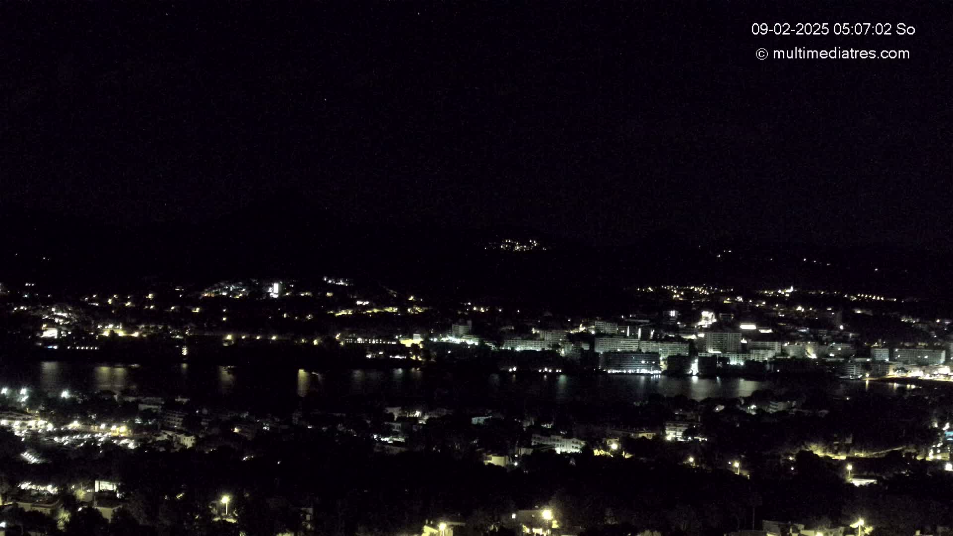 A nighttime, high-angle view shows a city and body of water illuminated by scattered lights under a clear, dark sky.