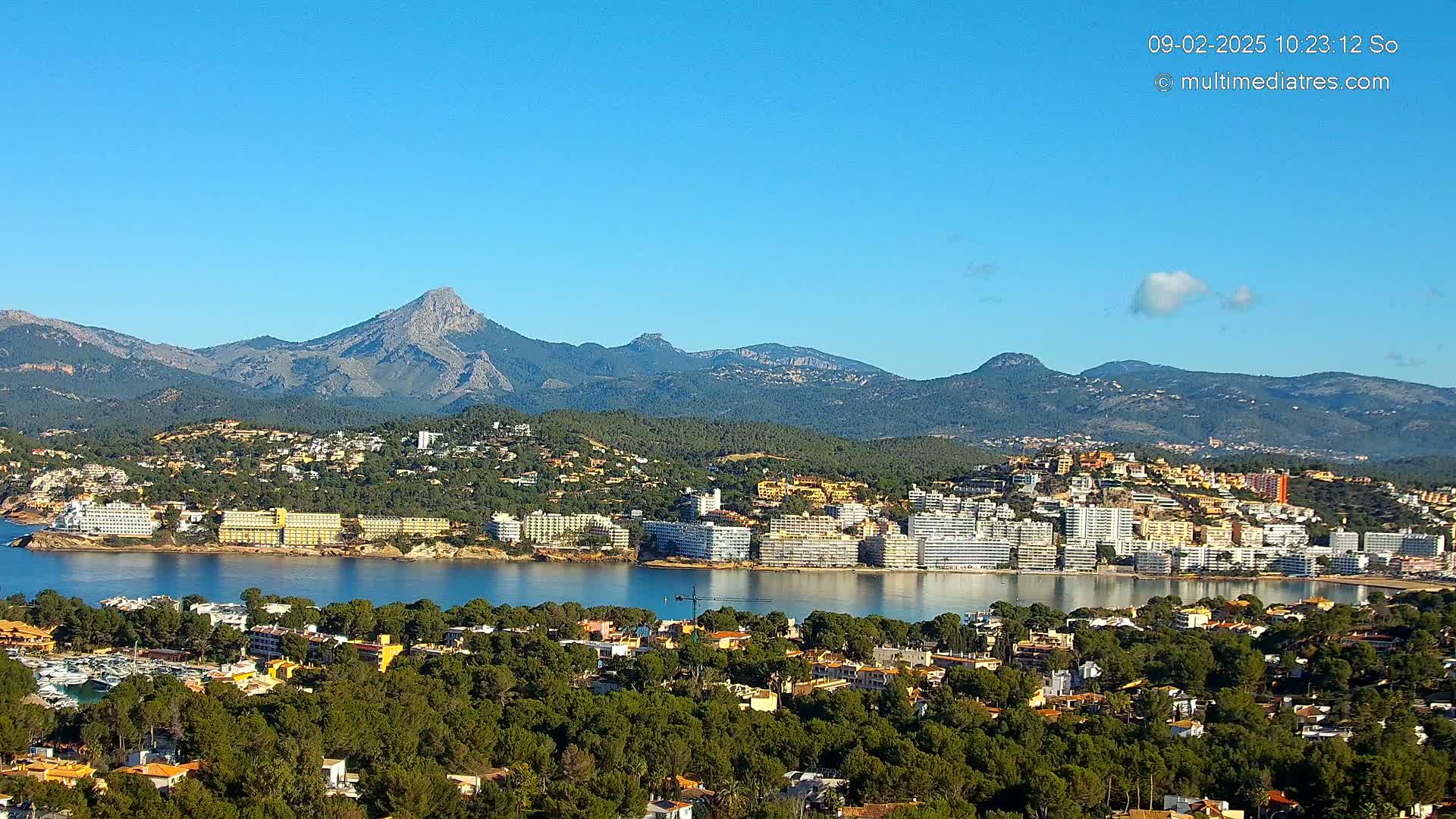A coastal town nestled in a bay, surrounded by lush green hills and mountains under a clear blue sky.