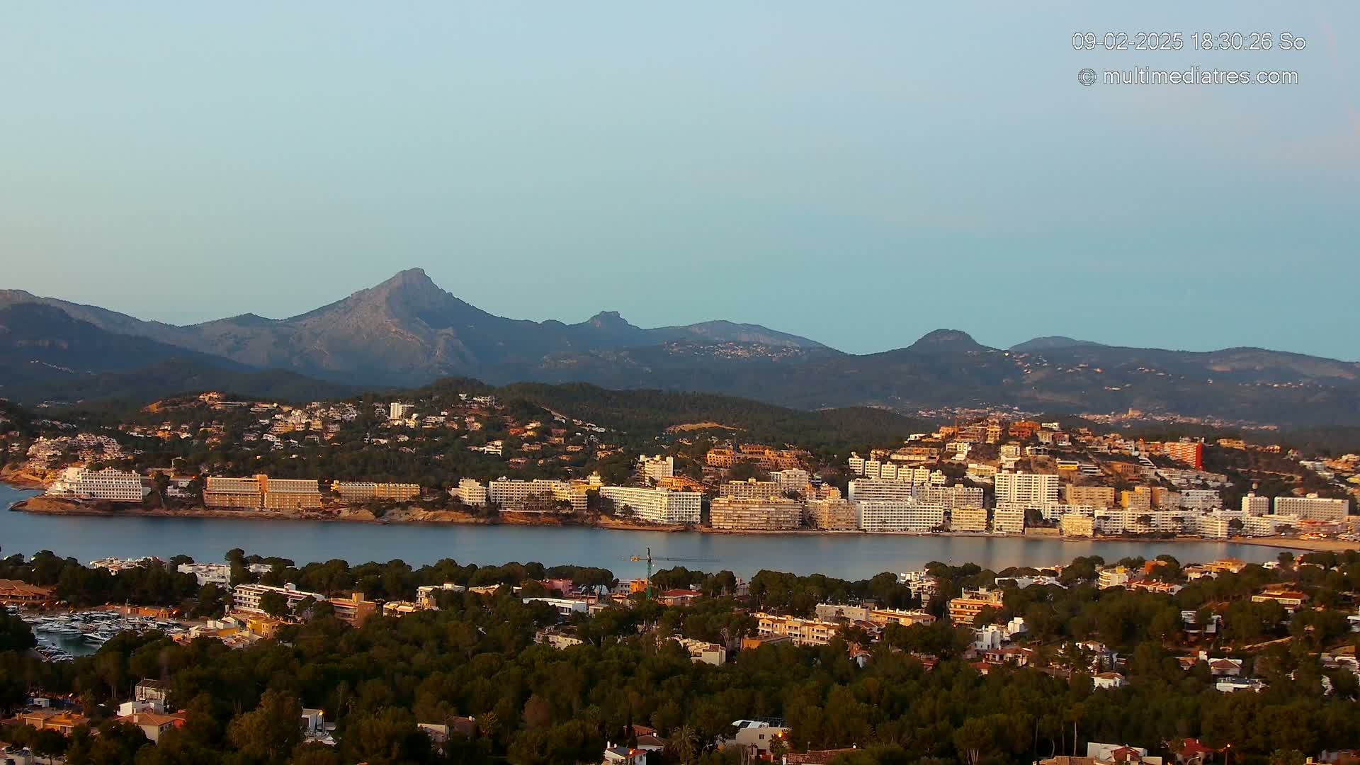 A coastal town nestled between a calm bay and a mountain range under a clear, twilight sky.