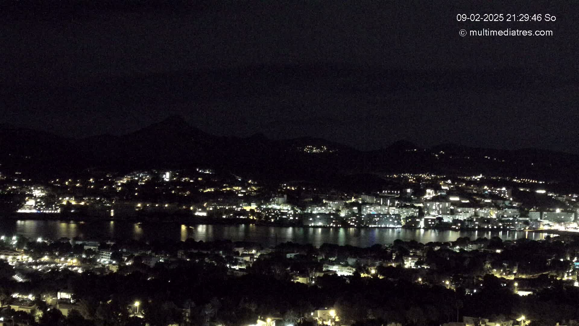 A nighttime view of a city nestled beside a body of water, illuminated by lights, with dark mountains in the background under a clear sky.