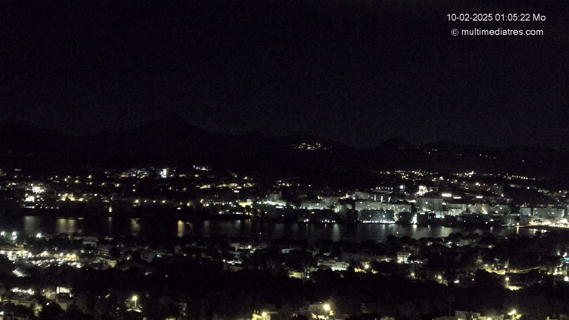 A nighttime, high-angle view shows a city and body of water surrounded by dark hills.