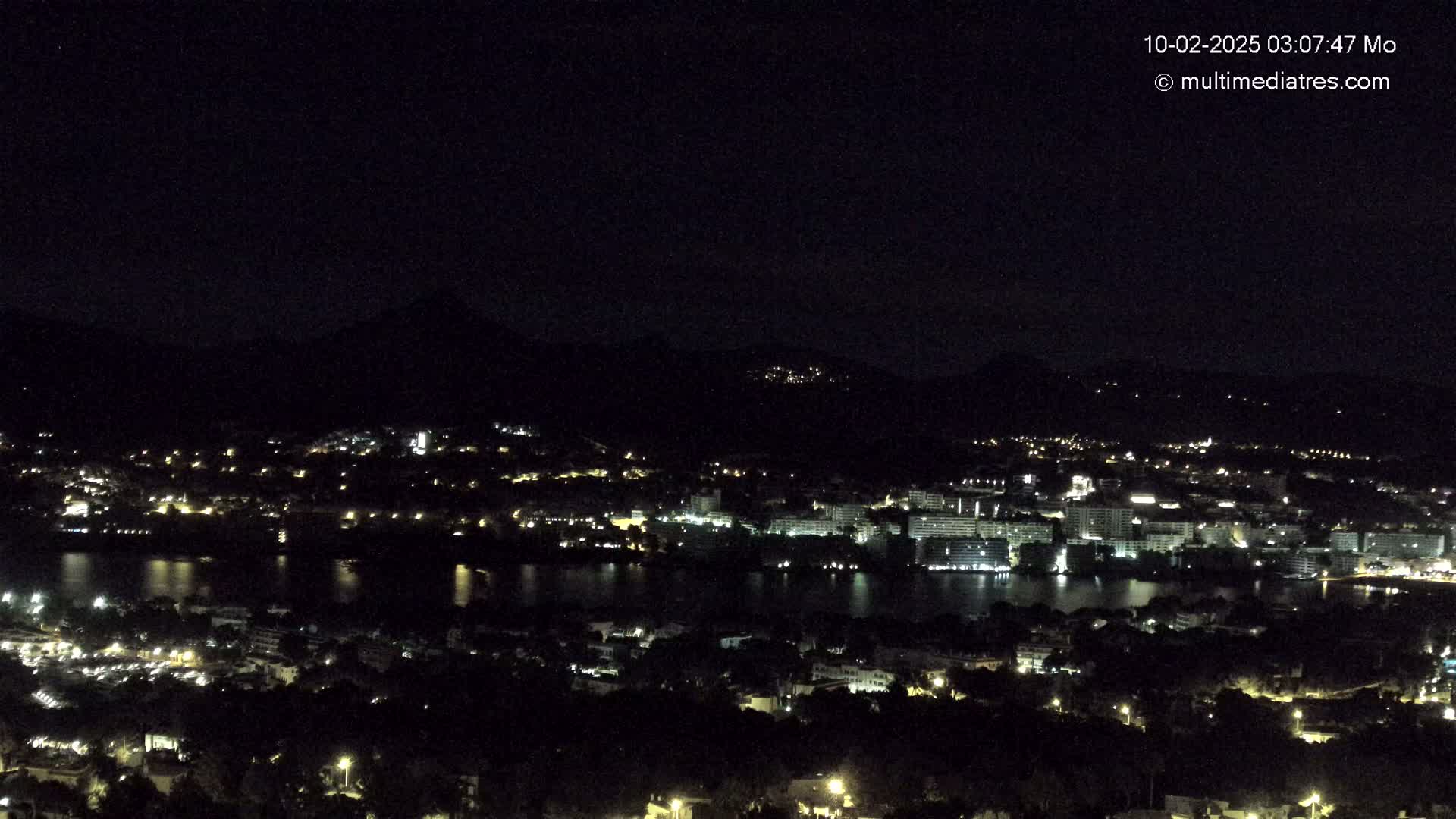 A nighttime, long-range view of a city with a body of water illuminated by scattered lights, nestled between dark hills under a clear, starless sky.