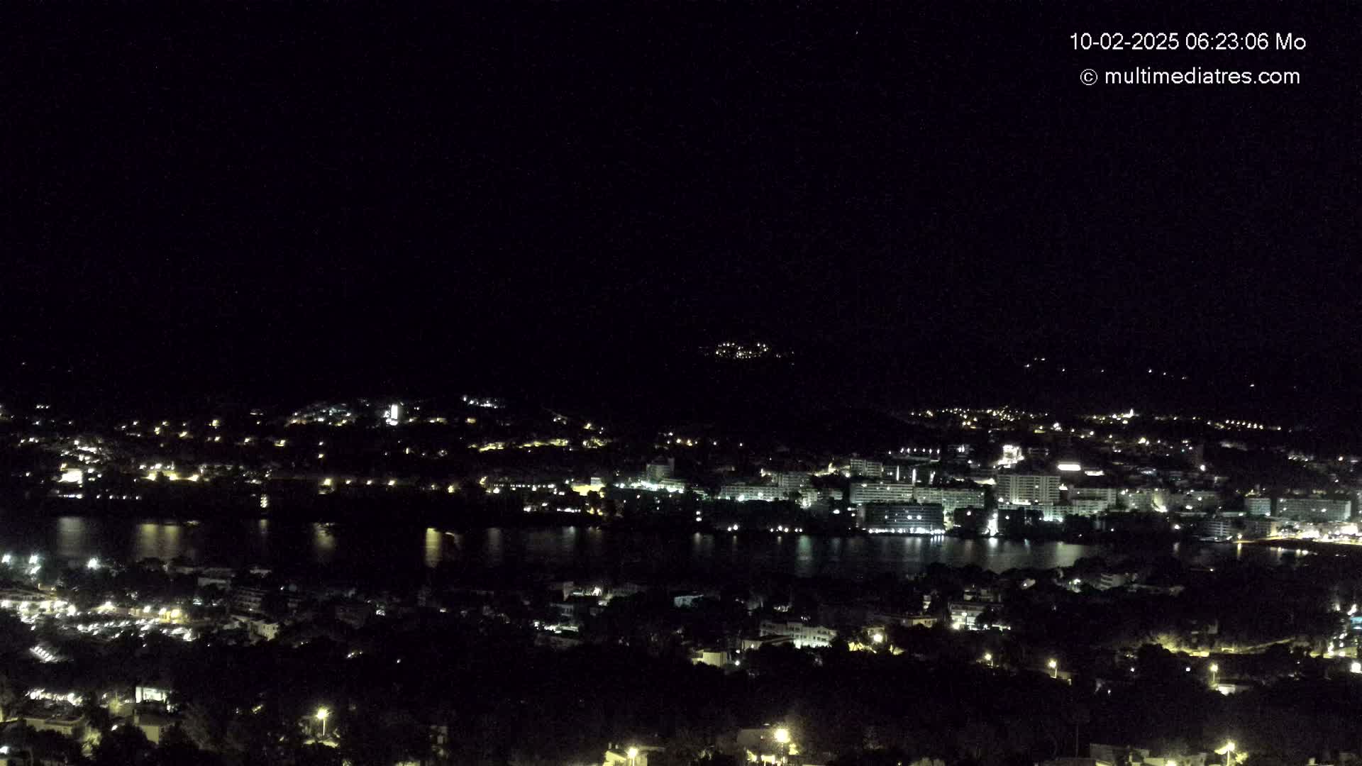 A nighttime, high-angle view shows a city with lights illuminating buildings near a body of water, all under a clear, dark sky.
