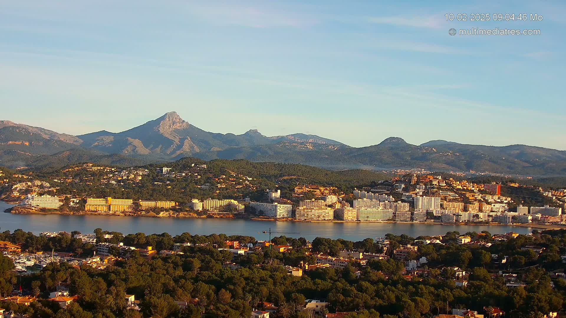 A coastal town nestled amongst green hills and mountains under a clear, bright sky.