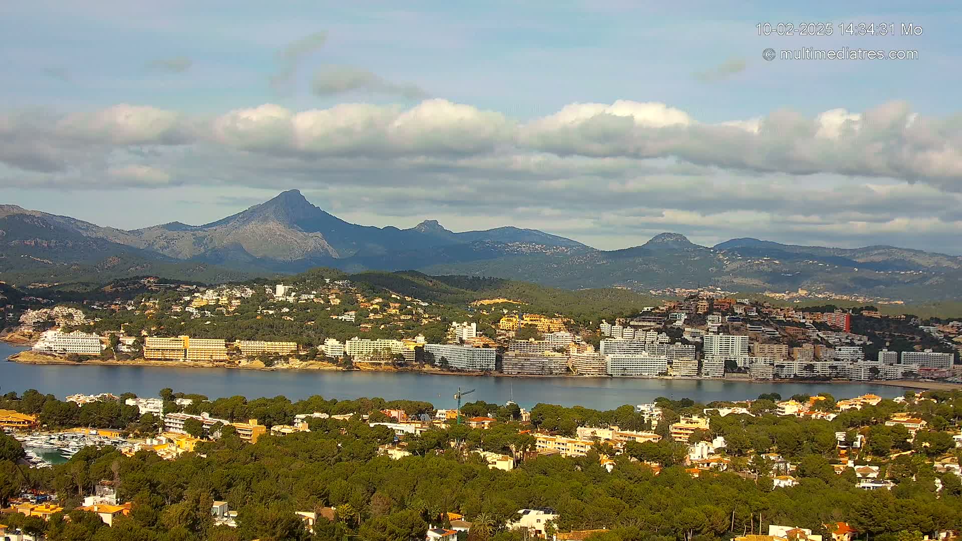 A coastal town nestled among green hills and mountains under a partly cloudy sky.