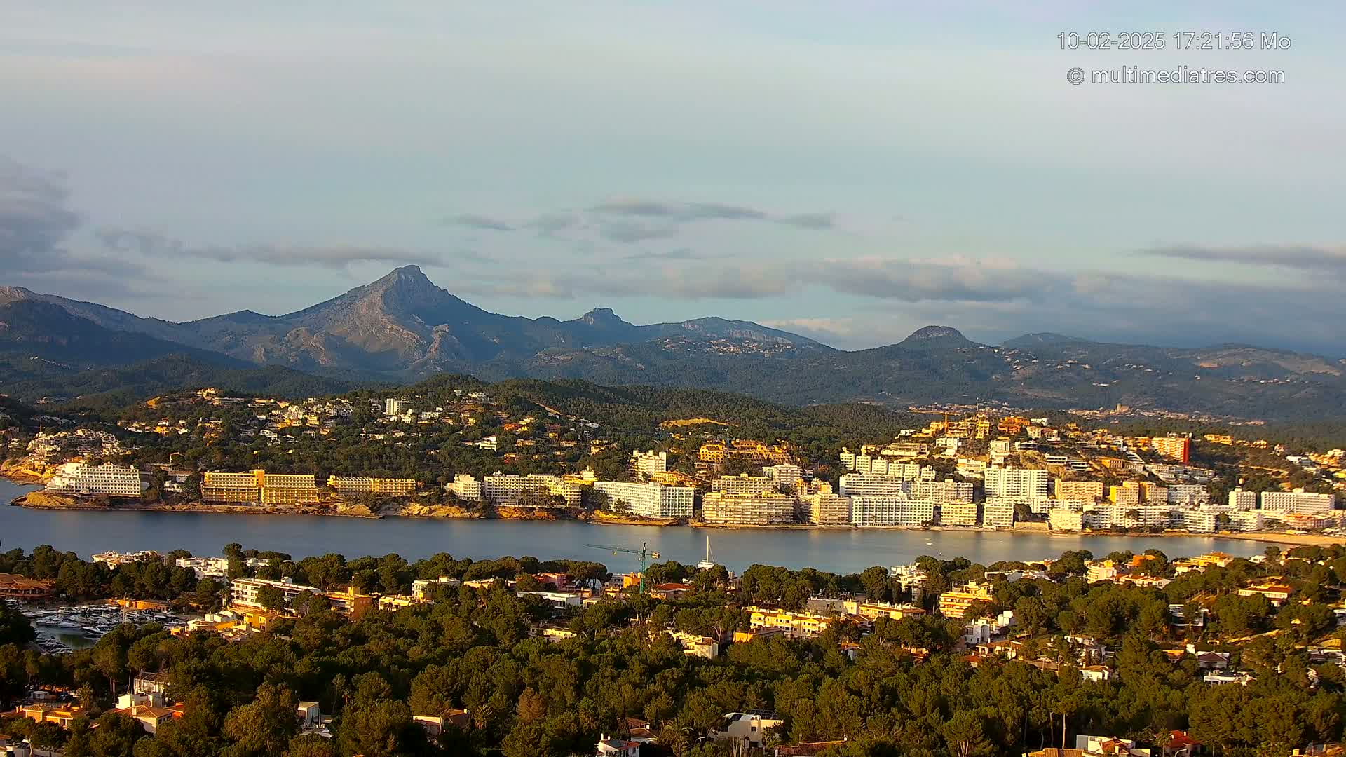 A coastal town nestled amongst green hills and mountains under a partly cloudy sky, with a calm body of water separating the town from a larger area of buildings.