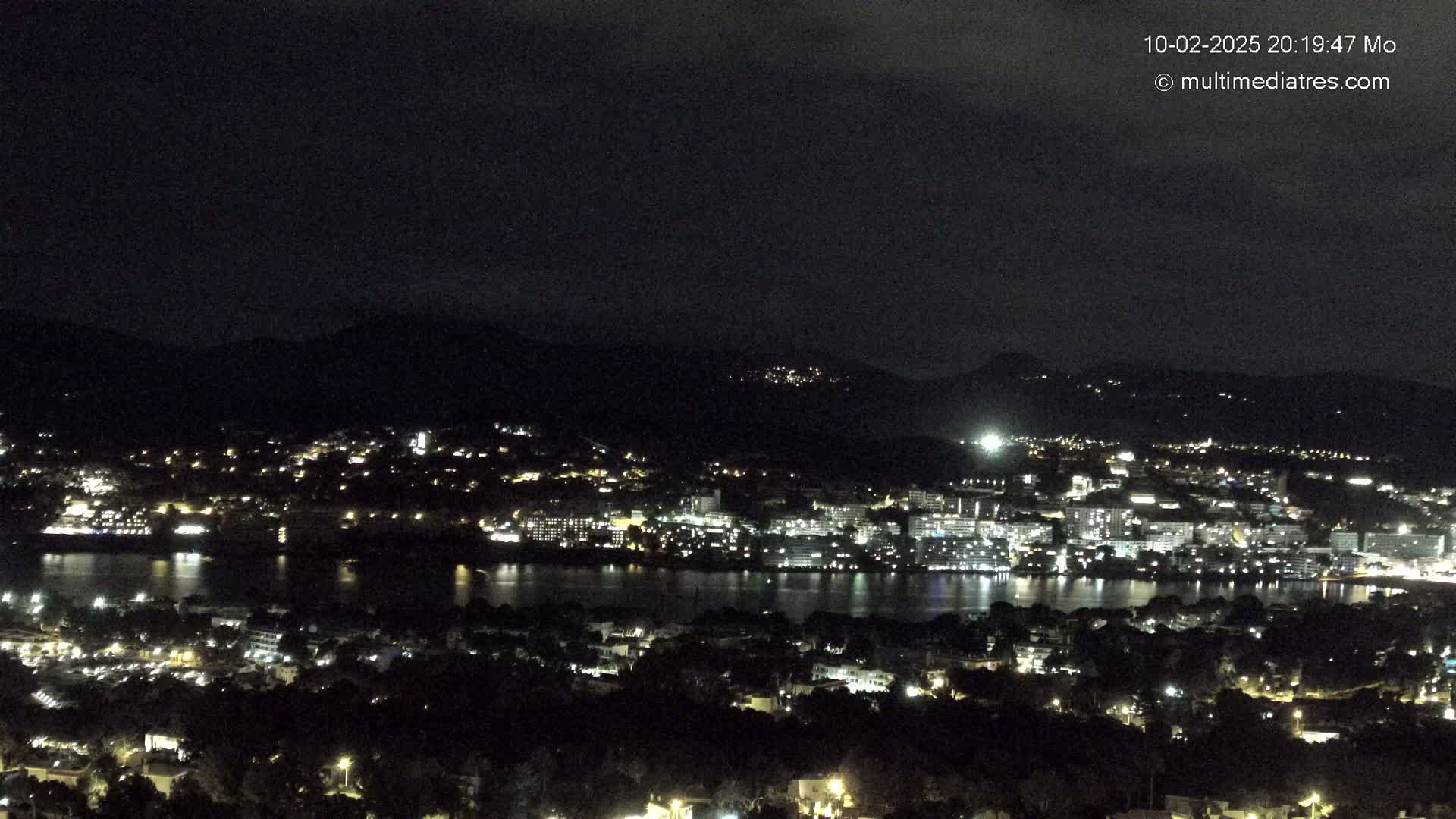 A nighttime, high-angle view of a coastal city illuminated by numerous lights, with a calm body of water separating it from a hillside of twinkling lights.