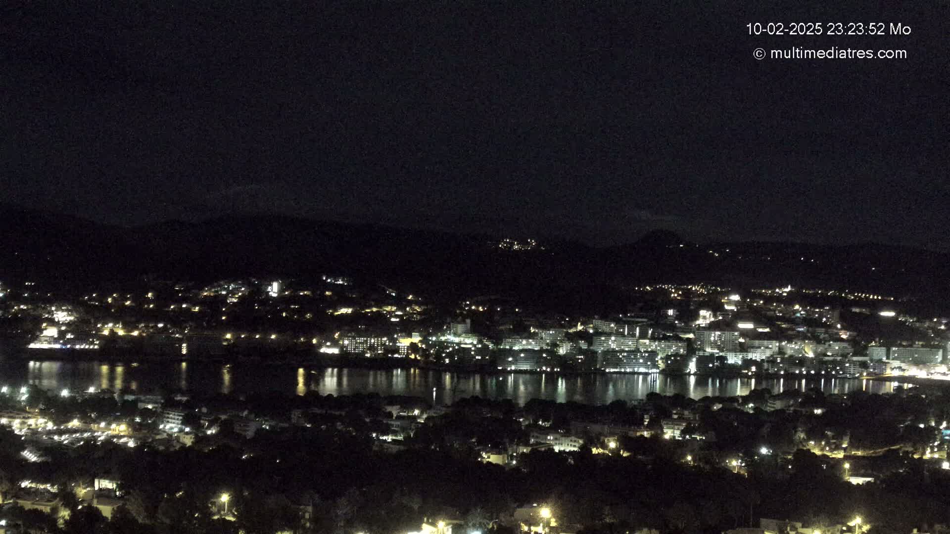A nighttime, high-angle view of a coastal town with its lights reflected in a calm body of water, nestled amongst dark hills under a clear, dark sky.