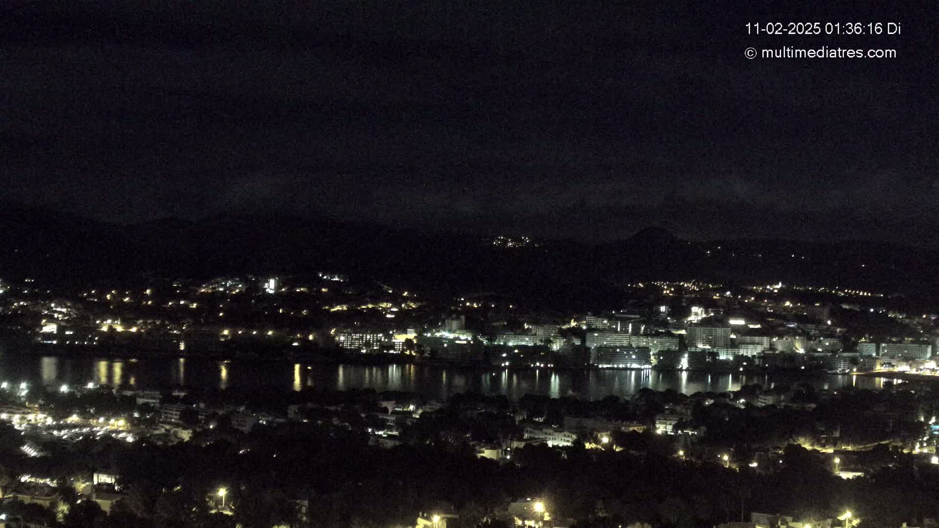 A nighttime view of a city with a body of water reflecting lights, situated near dark mountains under a clear, dark sky.