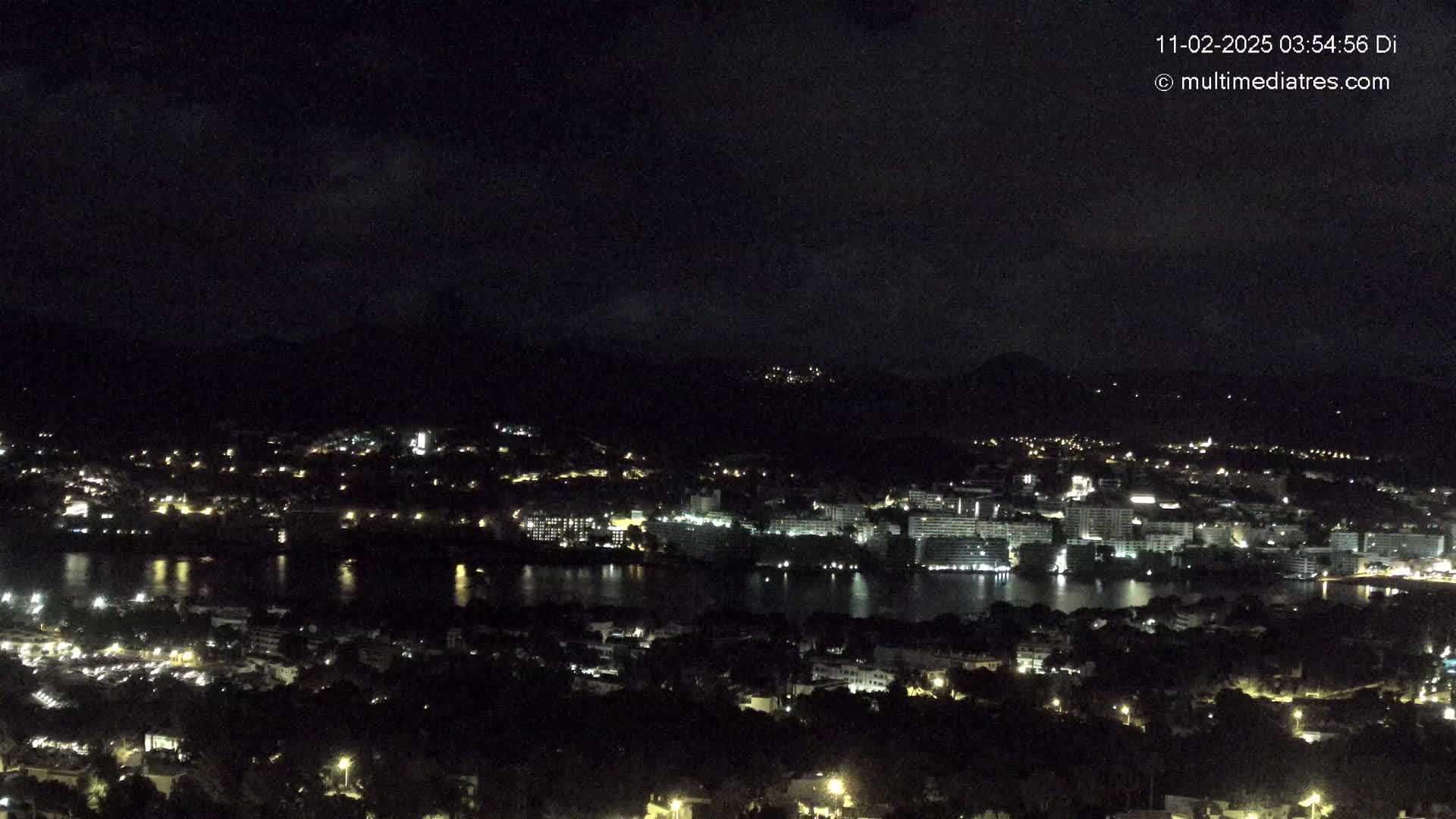 A nighttime view of a coastal town with many lights illuminating buildings near a dark body of water, under a mostly cloudy sky.