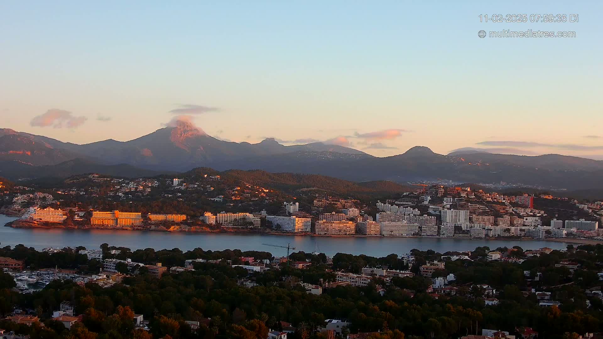 A coastal town nestled at the base of sunlit mountains, under a pale pink and blue sunrise sky.