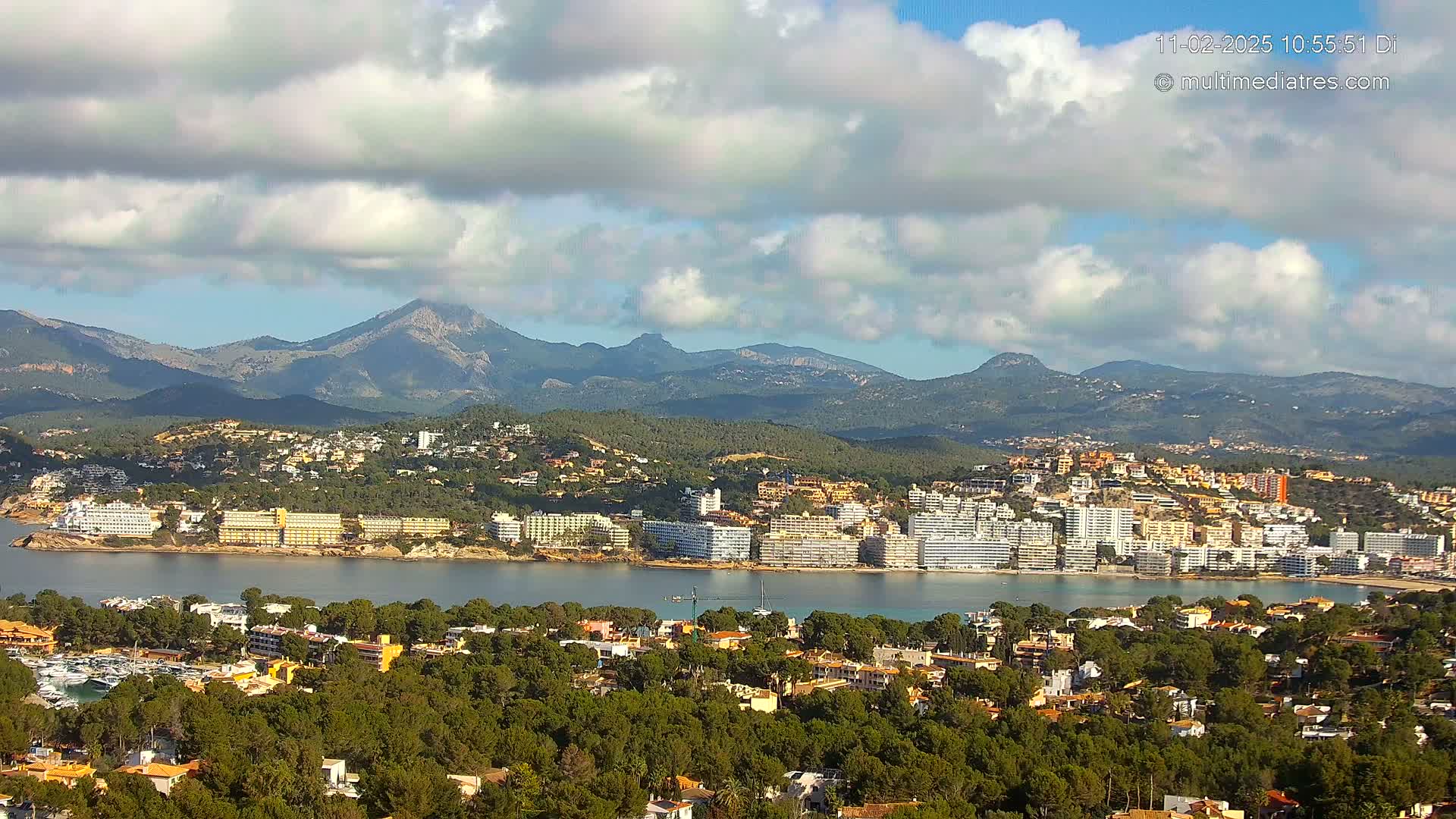 A coastal town nestled between a calm bay and a mountain range under a partly cloudy sky.