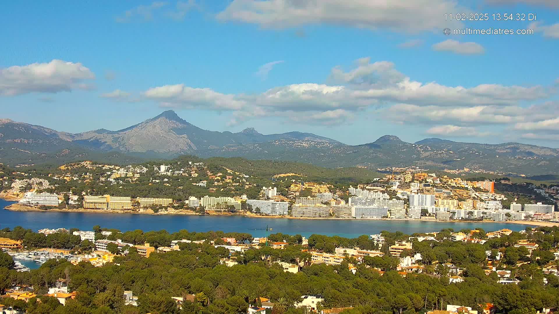 A coastal town nestled among green hills and mountains under a mostly sunny sky with scattered clouds.