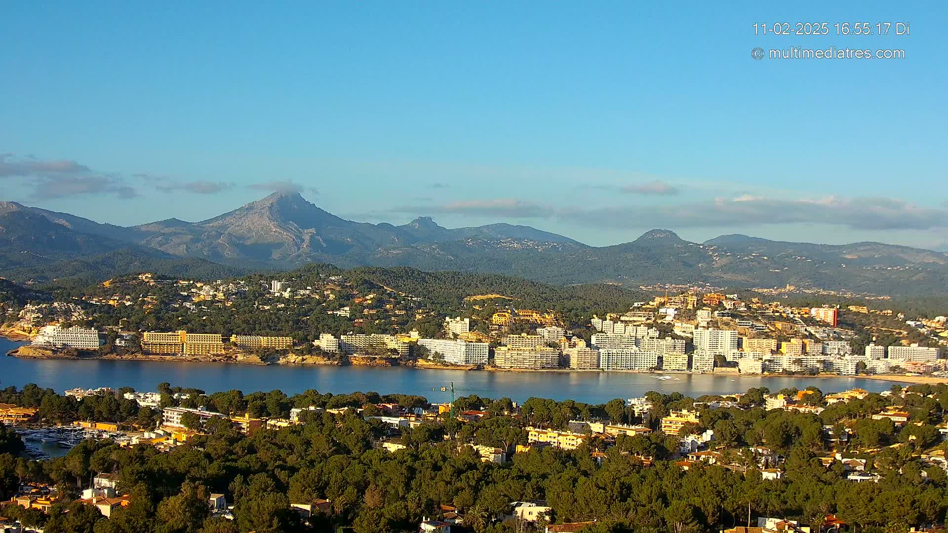 A coastal town nestled among green hills and mountains under a clear blue sky, with a calm body of water separating the town from a row of hotels.