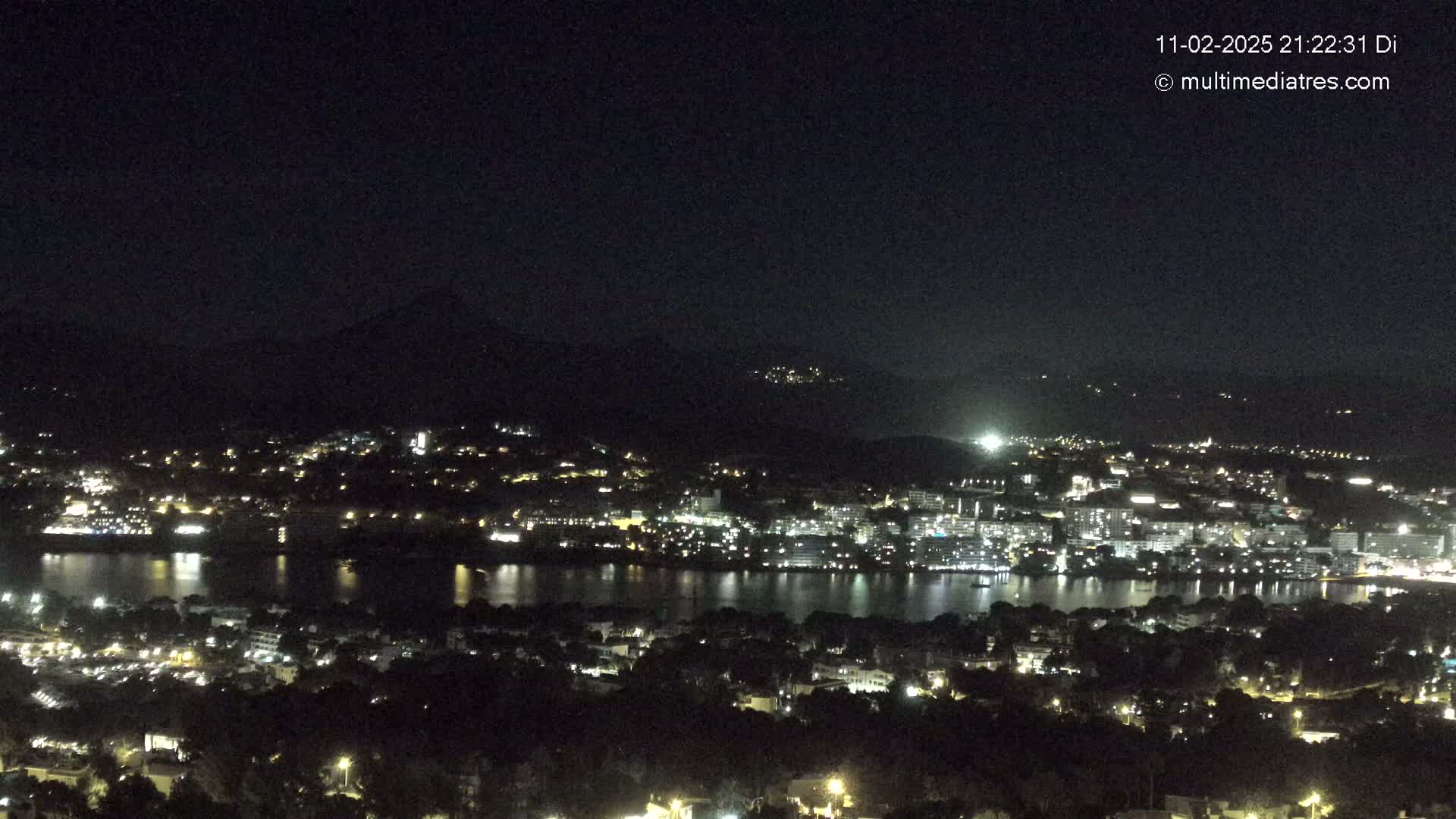 A nighttime view of a city and body of water, illuminated by lights, nestled against dark hills under a clear, dark sky.