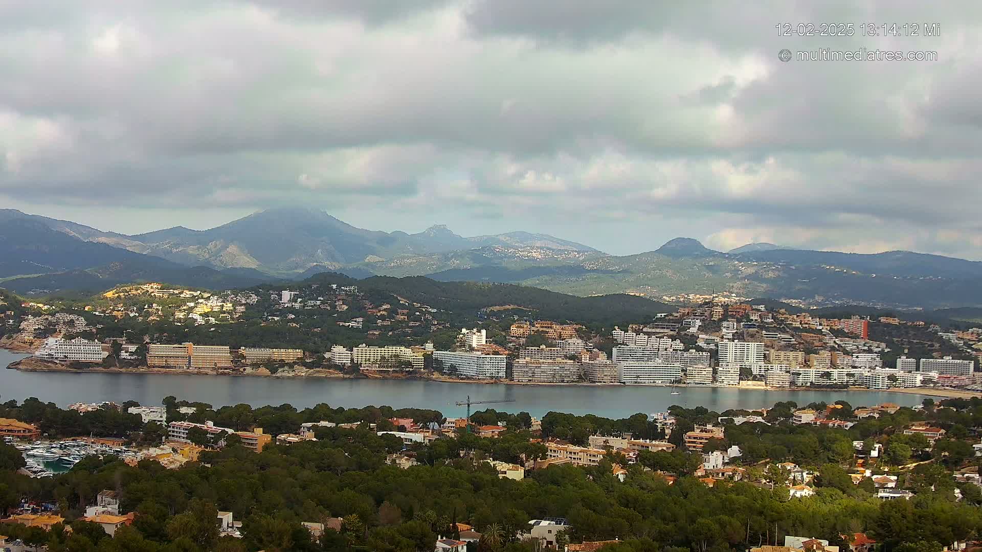 A coastal town nestled amongst green hills and mountains under a cloudy sky, with a calm bay separating the town from a larger waterfront area.