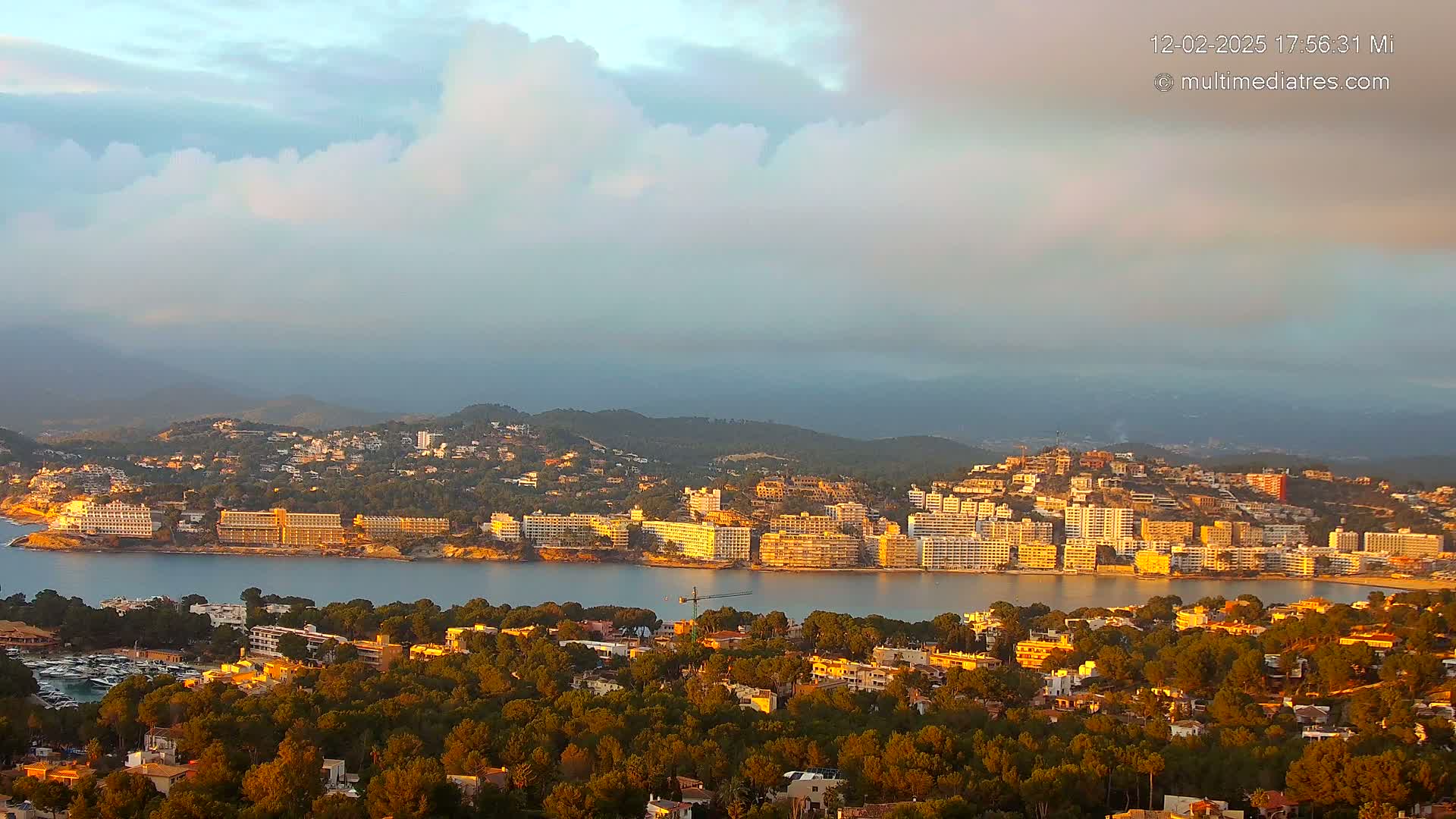 A coastal town nestled in a bay, surrounded by hills and partly cloudy skies, is bathed in the golden light of sunrise or sunset.