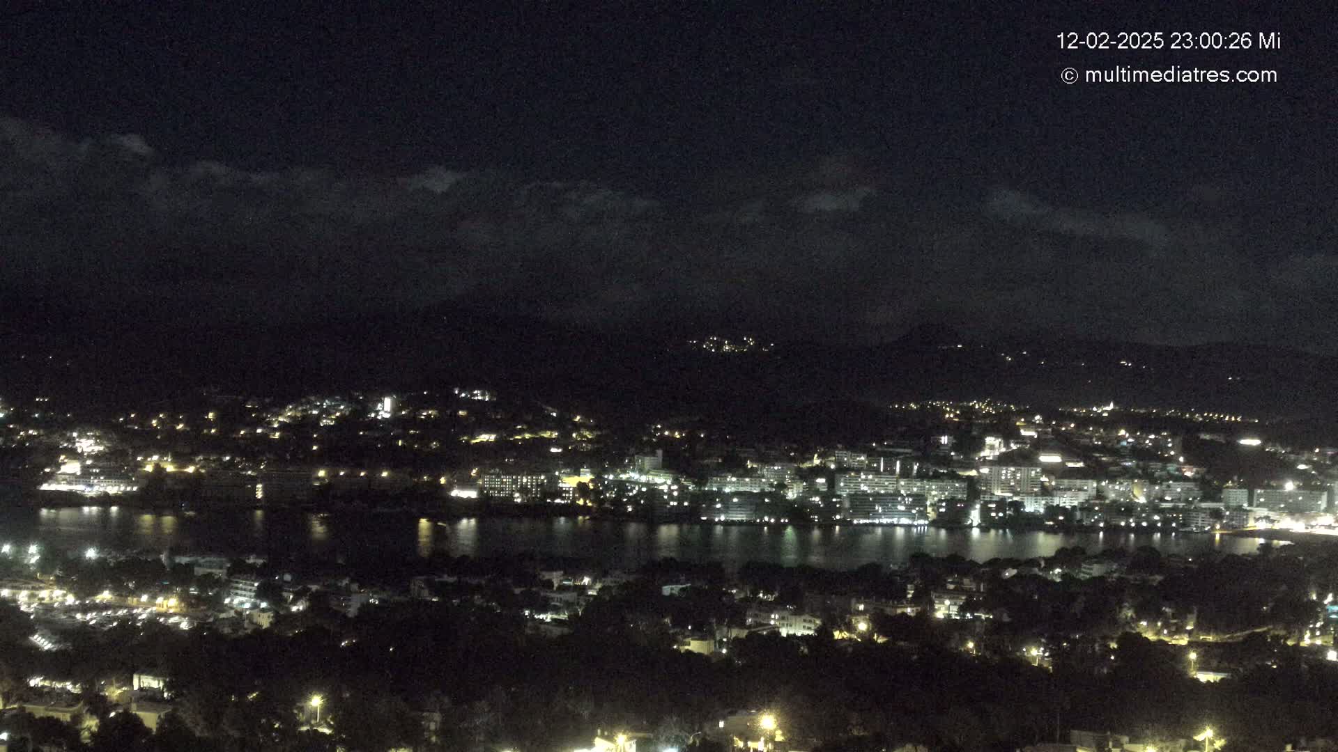 A nighttime view of a city illuminated with lights, situated beside a body of water, under a mostly clear, dark sky.