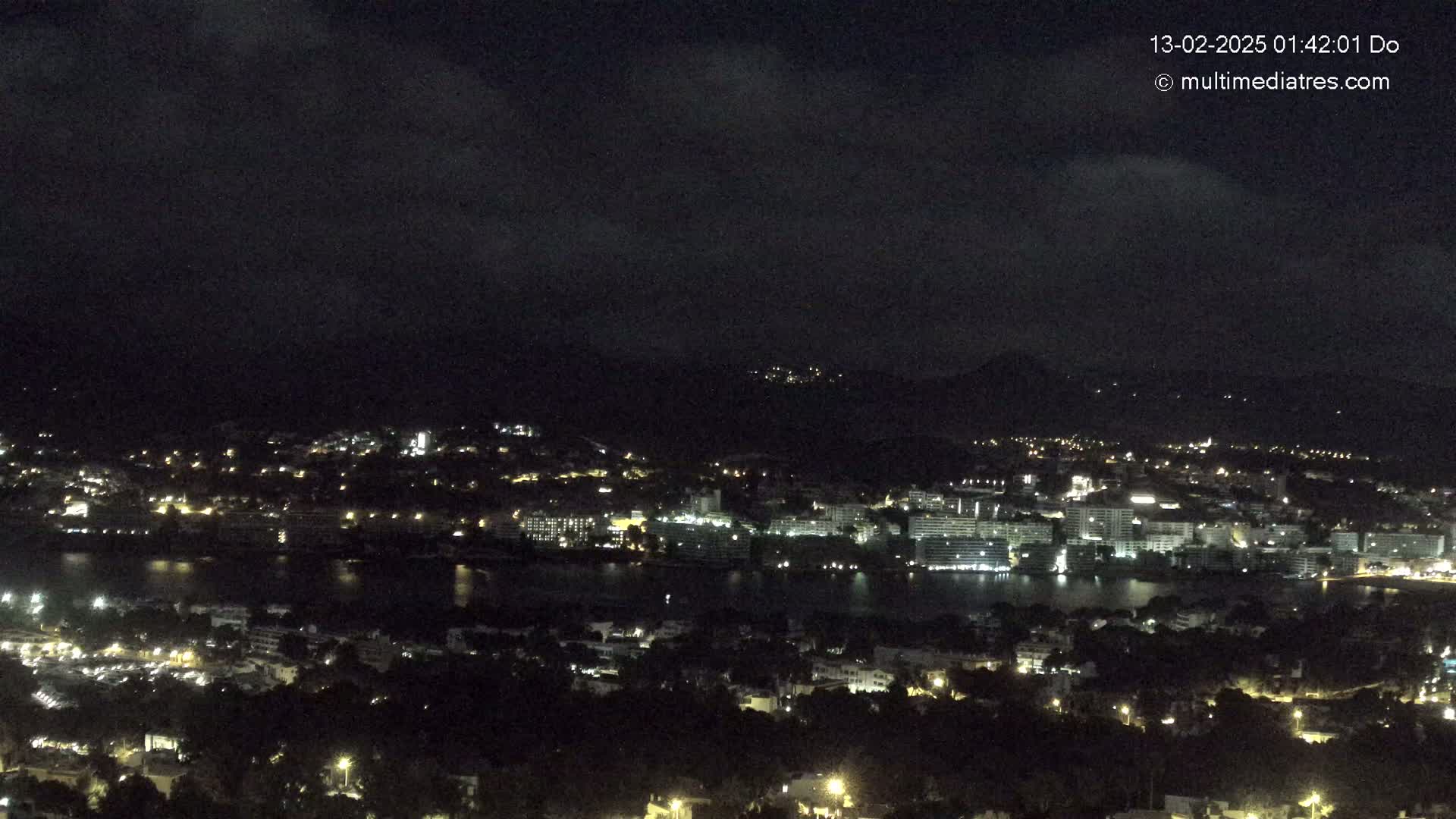 A nighttime, high-angle view shows a coastal town with buildings lit up along a dark body of water, under a mostly cloudy sky.