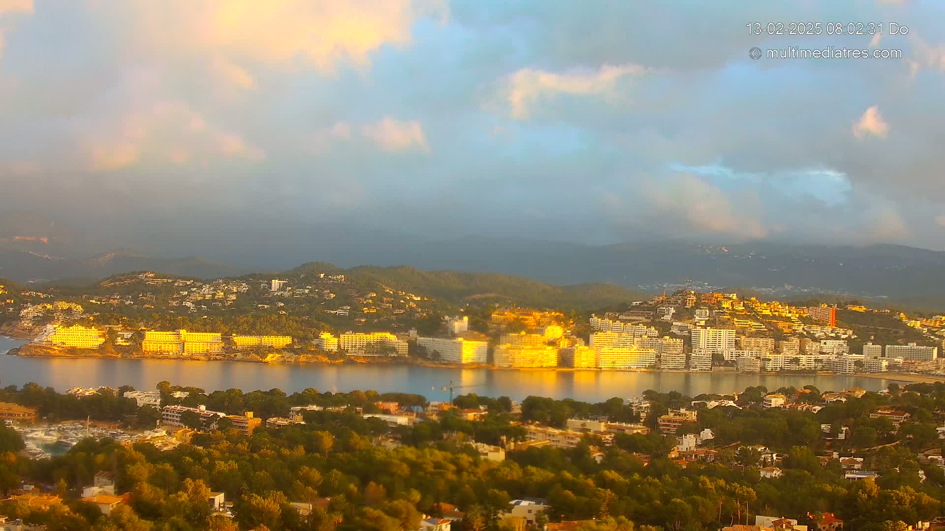 A coastal town nestled among hills is bathed in the golden light of sunrise under a partly cloudy sky.