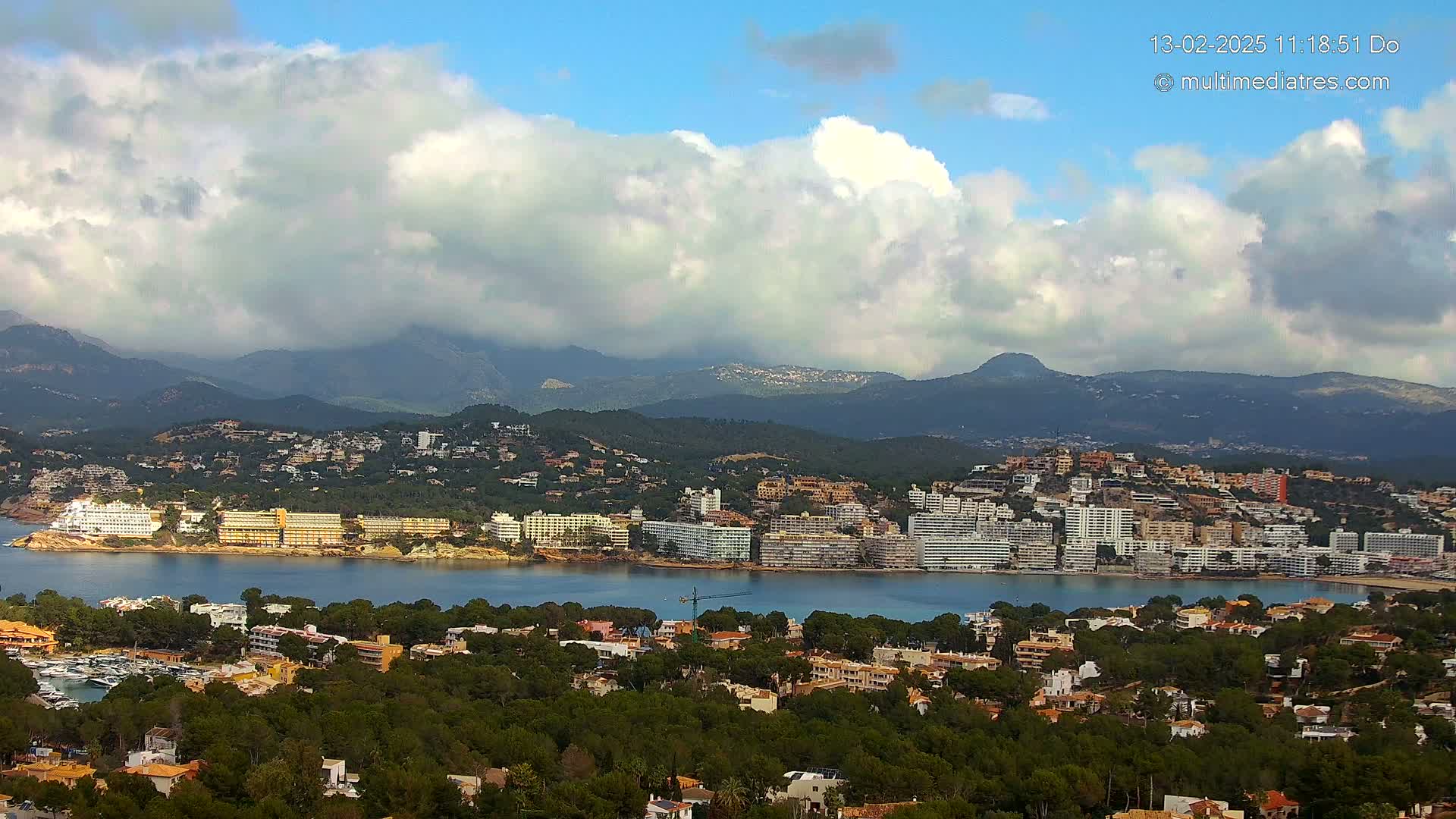 A coastal town nestled between a calm bay and mountainous terrain under a partly cloudy sky.