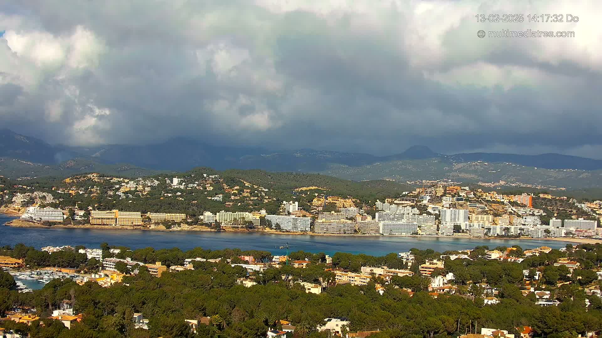 A coastal town nestled among green hills and mountains under a partly cloudy sky, with a calm body of water separating the town from a line of hotels.