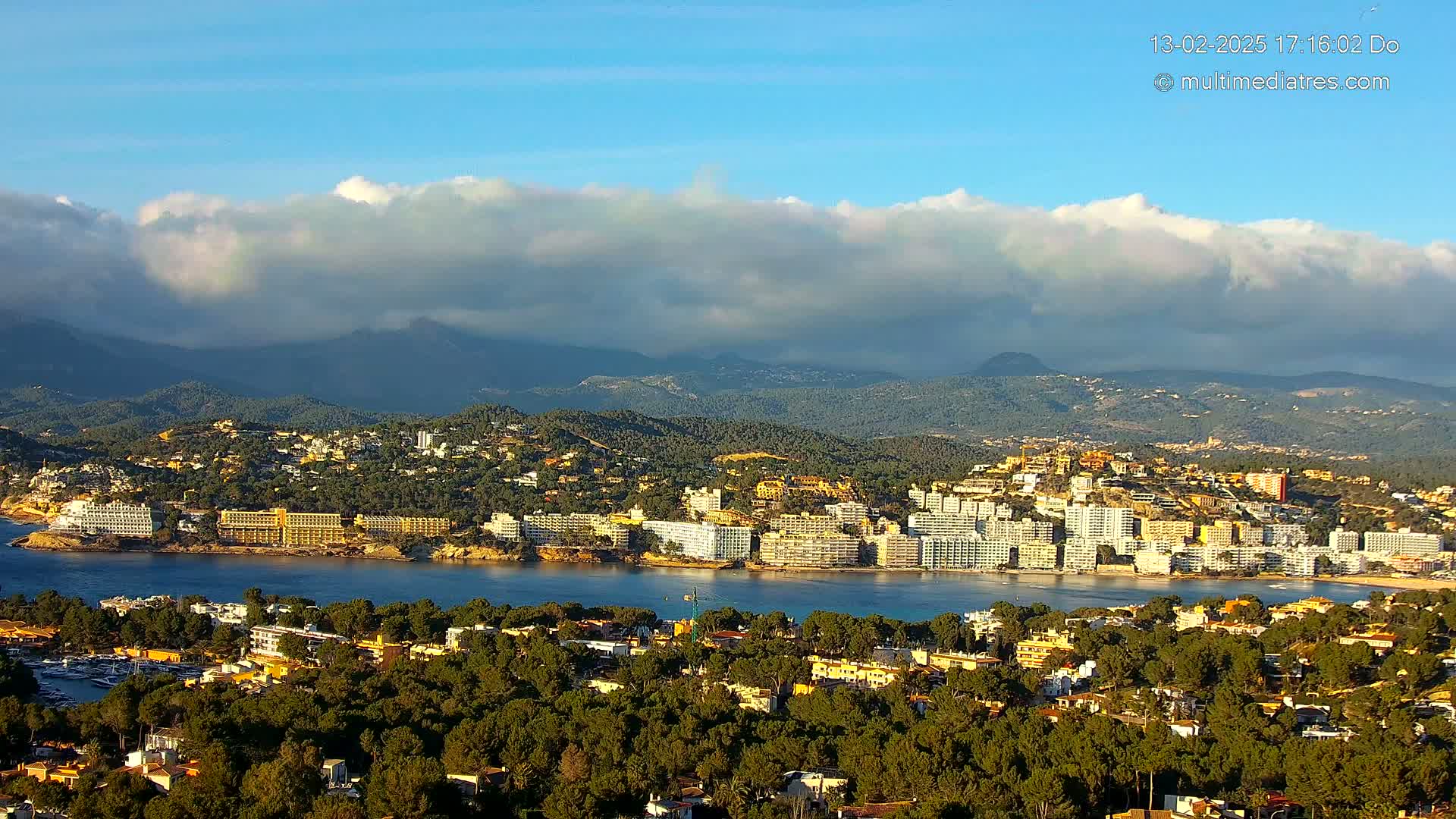 A coastal town nestled among green hills and mountains under a partly cloudy sky, with a calm body of water separating the town from a line of hotels.