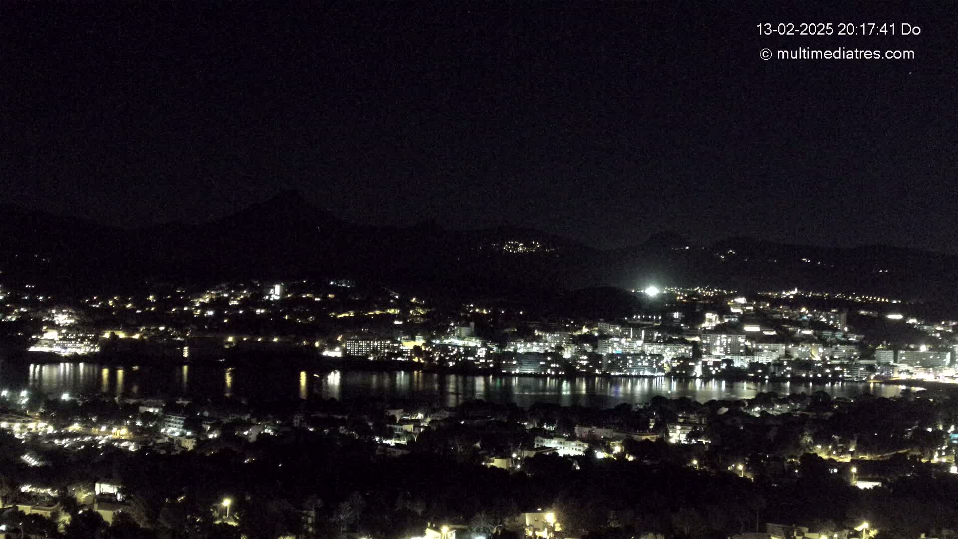 A nighttime view of a city illuminated by lights, situated beside a body of water, with dark hills in the background under a clear, dark sky.