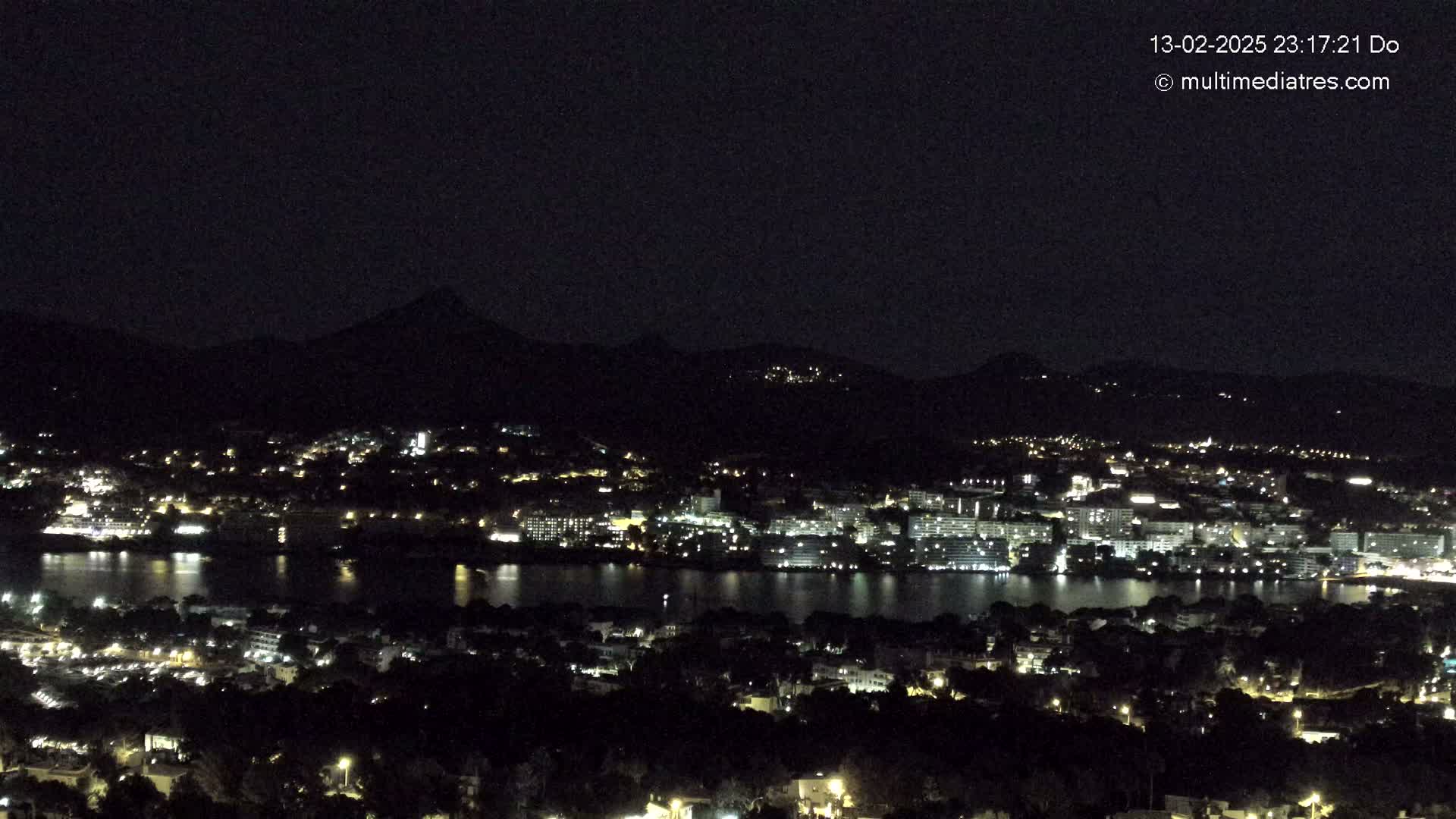 A nighttime view of a coastal city illuminated by lights, situated at the base of dark mountains under a clear, dark sky.
