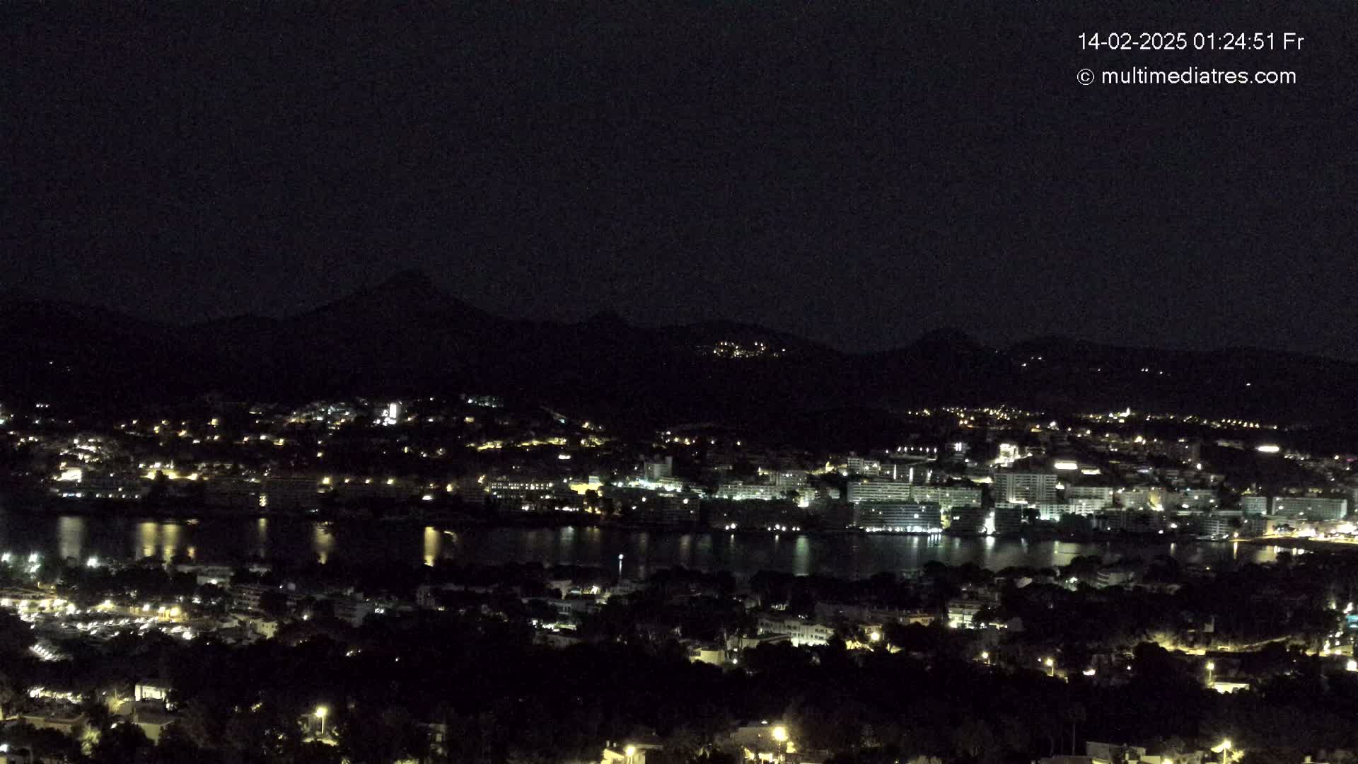 A nighttime view of a coastal town with numerous lights, situated on a body of water and surrounded by dark hills under a clear, dark sky.