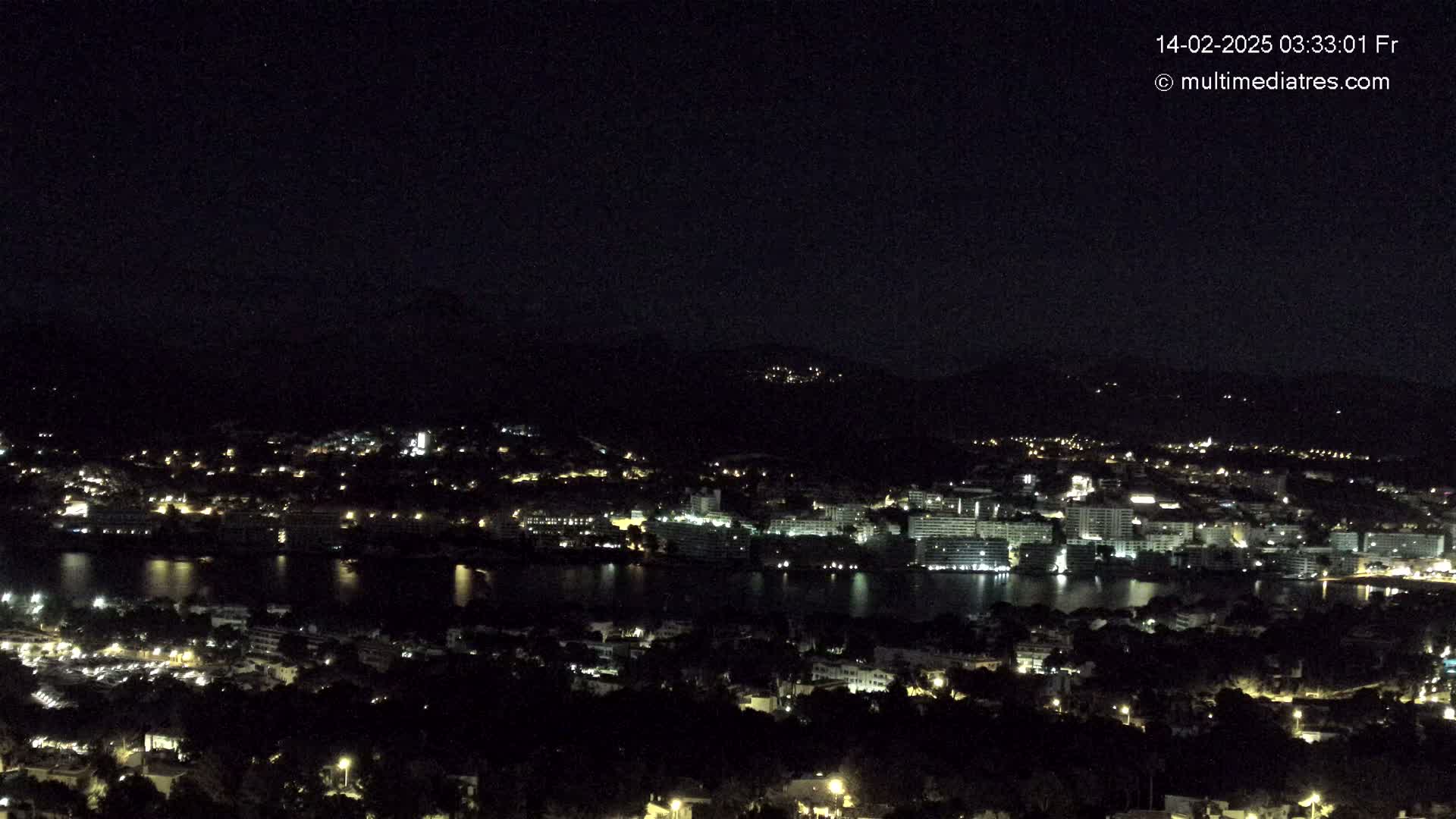 A nighttime, long shot of a city sprawling along a dark body of water, nestled between hills under a clear, dark sky.