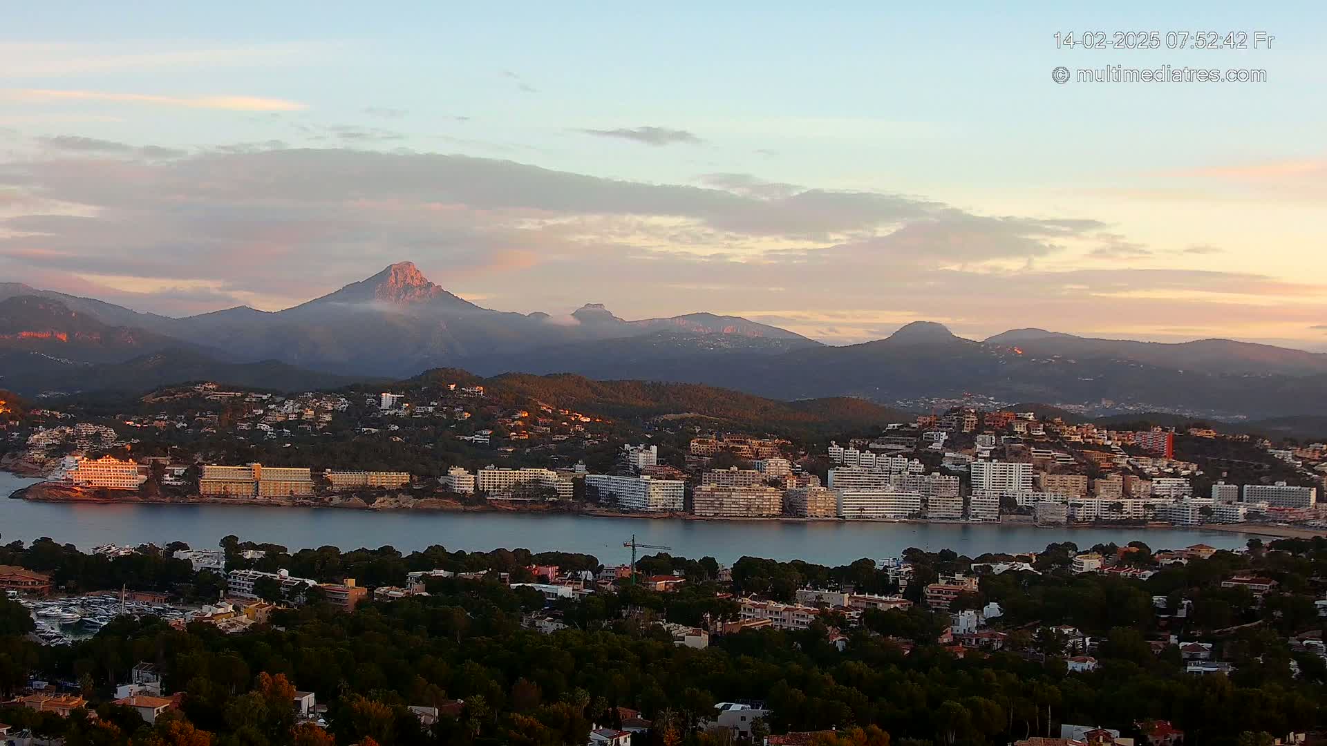 A coastal town nestled between a calm bay and a mountain range under a soft, pastel sunrise sky.