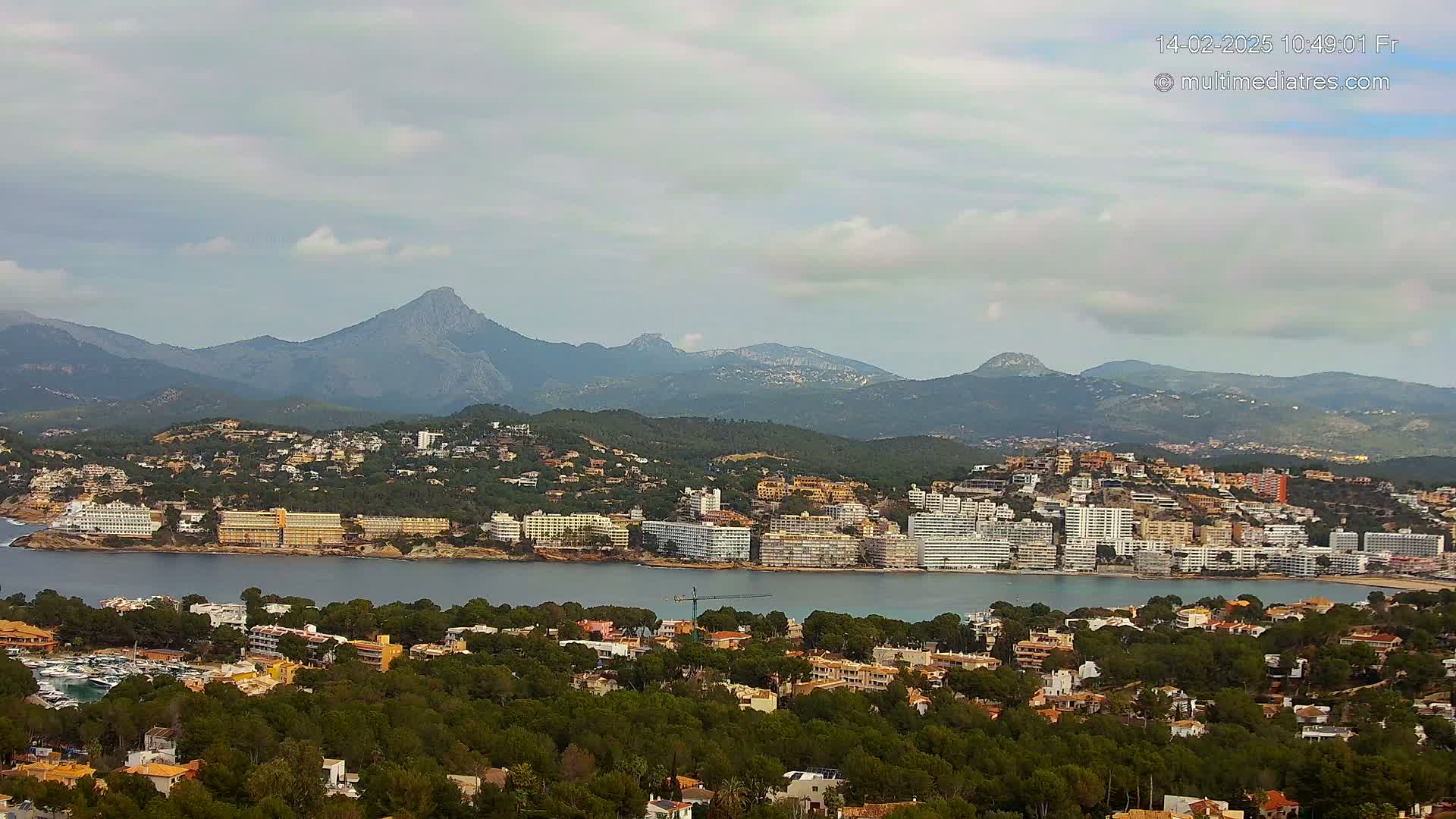 A coastal town nestled among green hills and mountains under a mostly cloudy sky overlooks a calm bay.