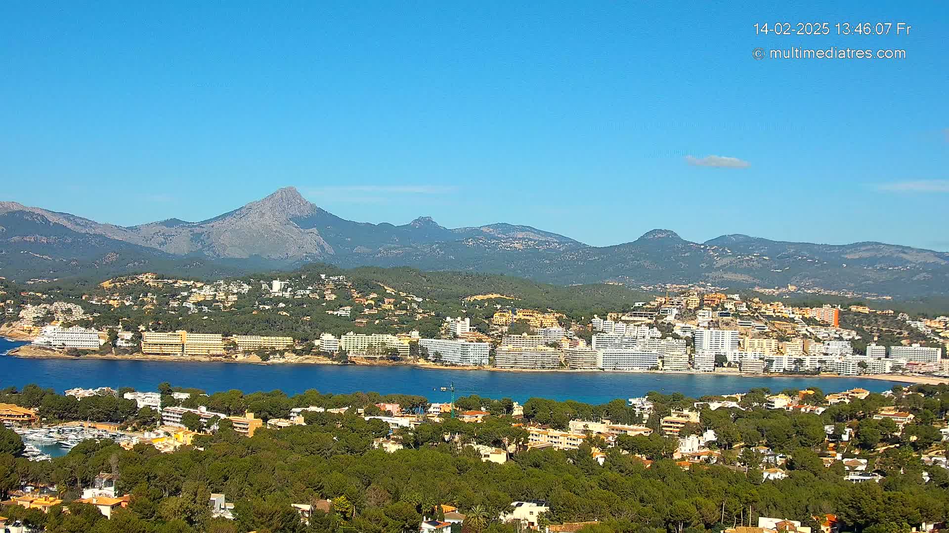A coastal town nestled among lush green hills and mountains under a clear blue sky, with a calm body of water separating the town from a row of hotels.