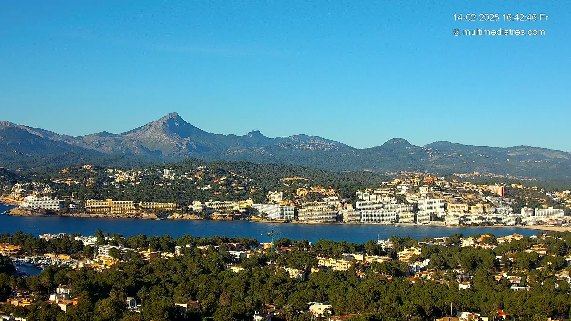 A coastal town nestled between a calm bay and a mountain range under a clear blue sky.