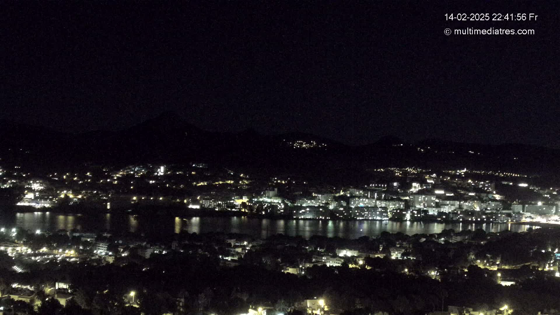 A nighttime view of a city illuminated by lights, situated beside a body of water, with dark hills rising in the background under a clear, dark sky.