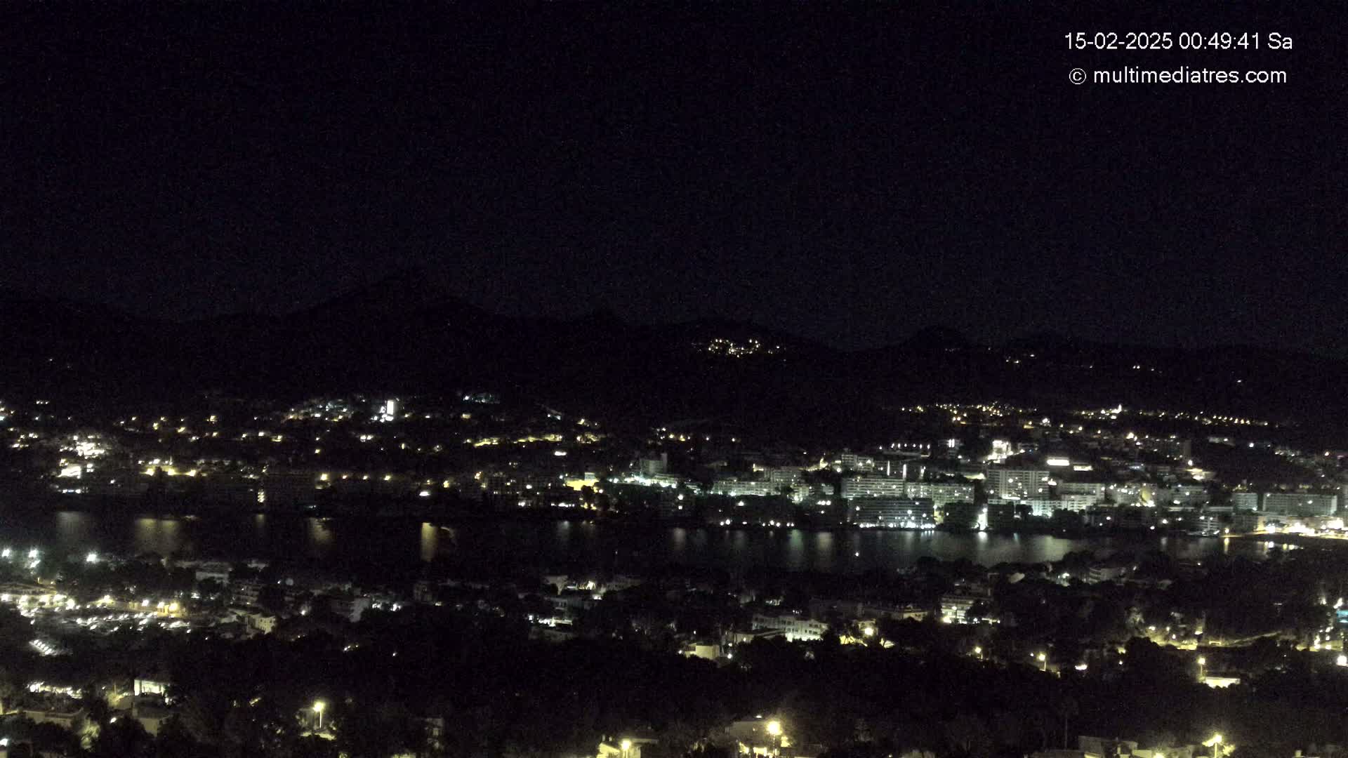 A nighttime view of a city nestled beside a body of water, illuminated by numerous lights, with dark hills rising in the background under a clear, dark sky.