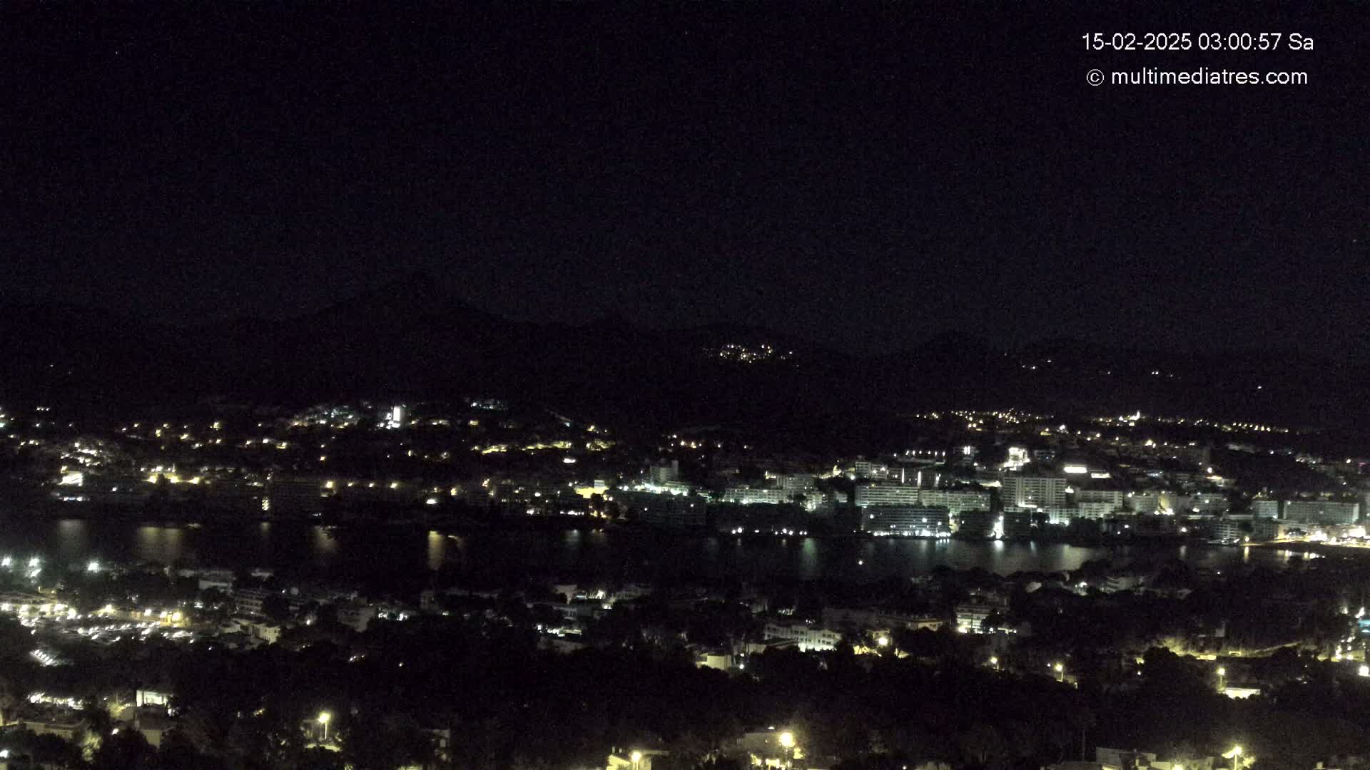 A nighttime, high-angle view shows a city with many lights nestled beside a dark body of water, all situated at the base of a dark mountain range under a clear, starless sky.